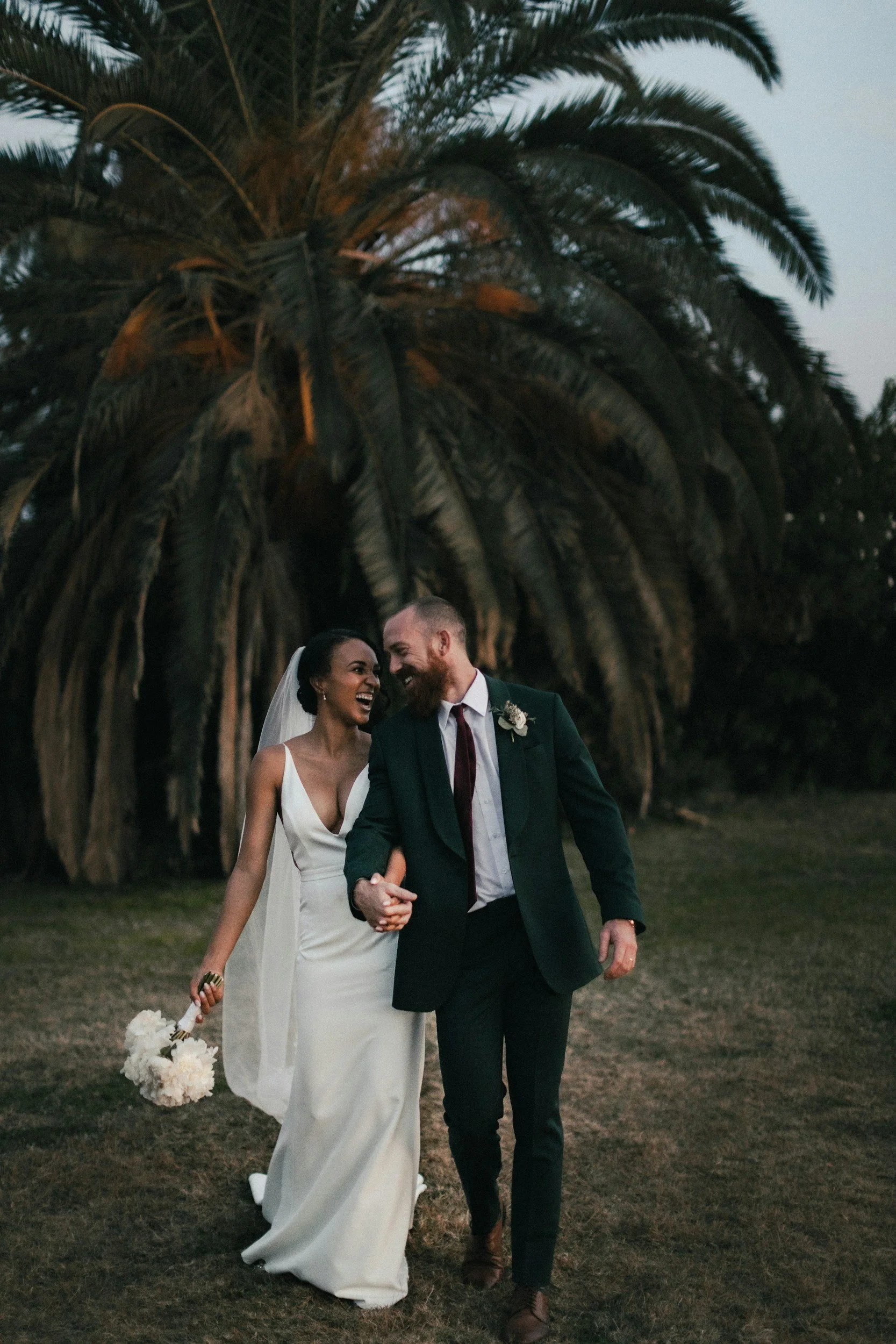 A newlywed couple holding hands and laughing outdoors at sunset, with a large palm tree in the background.