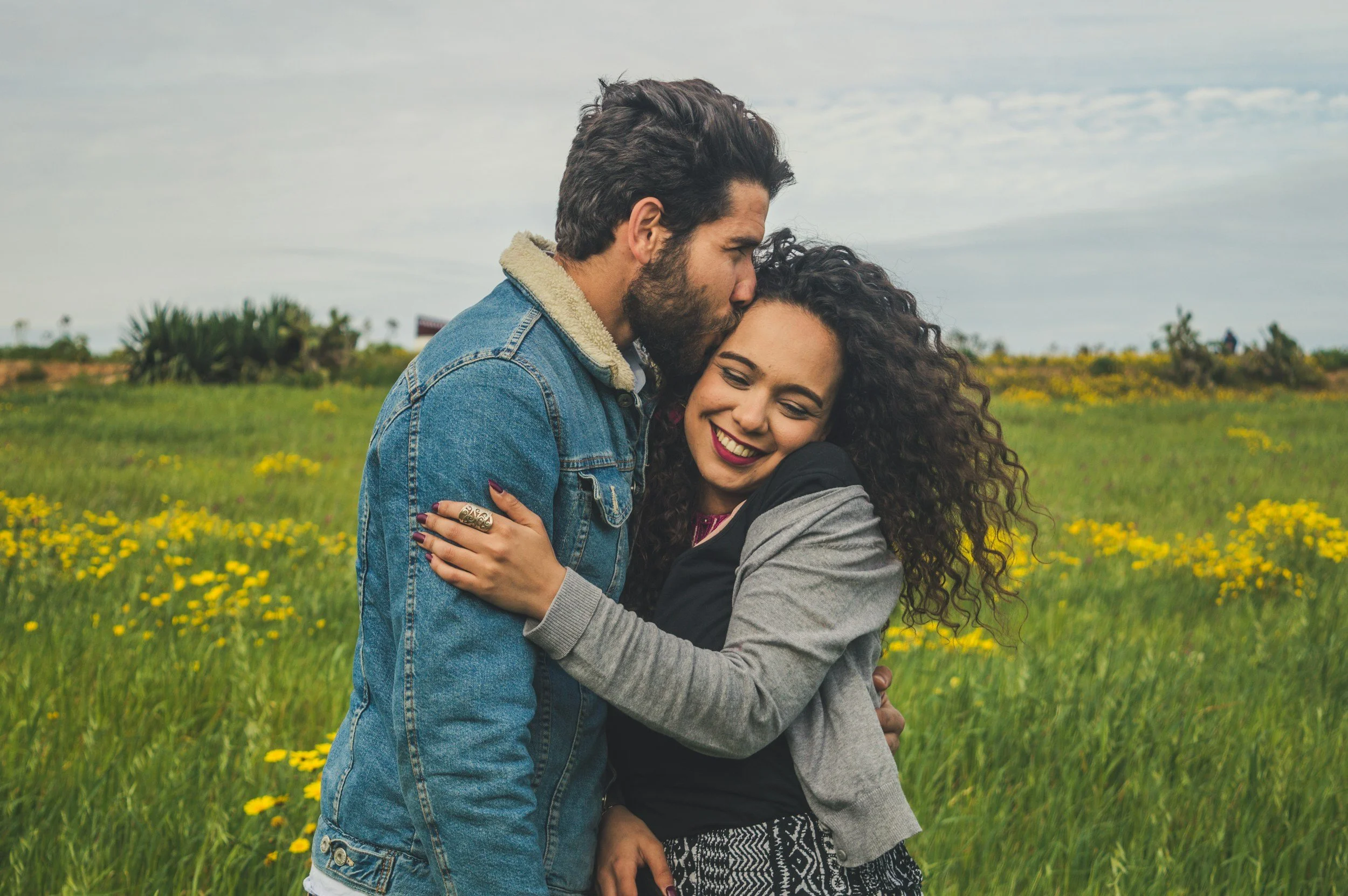 A young couple hugging in a grassy field with yellow flowers, the man is kissing the woman's forehead and she is smiling with her eyes closed.