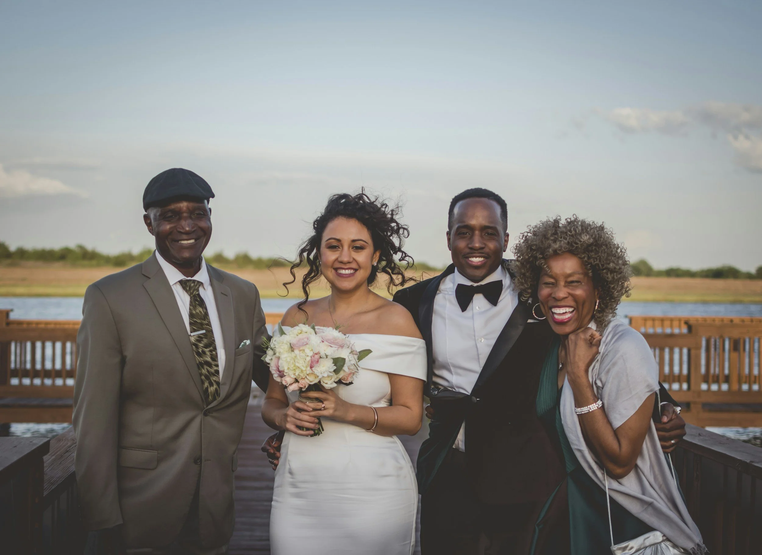 Group of four people at a wedding, including a bride in a white dress holding a bouquet, and three older adults, standing outdoors near water with a wooden railing in the background, smiling.