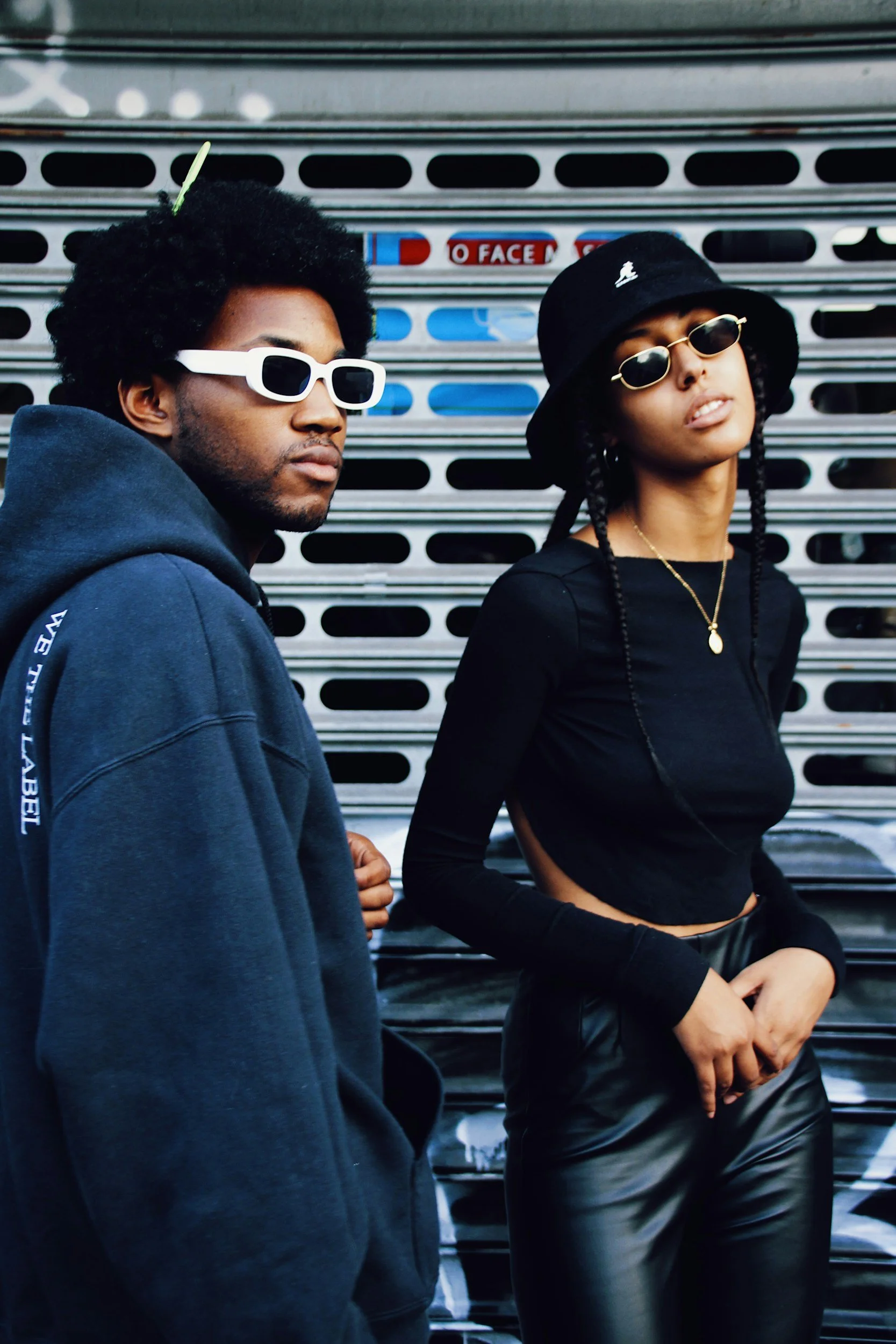 A man and woman standing in front of a metal roller shutter door, both wearing dark clothing and sunglasses. The man has curly hair with a hairpin and wears a hooded sweatshirt, while the woman has braids, a bucket hat, and a gold necklace.