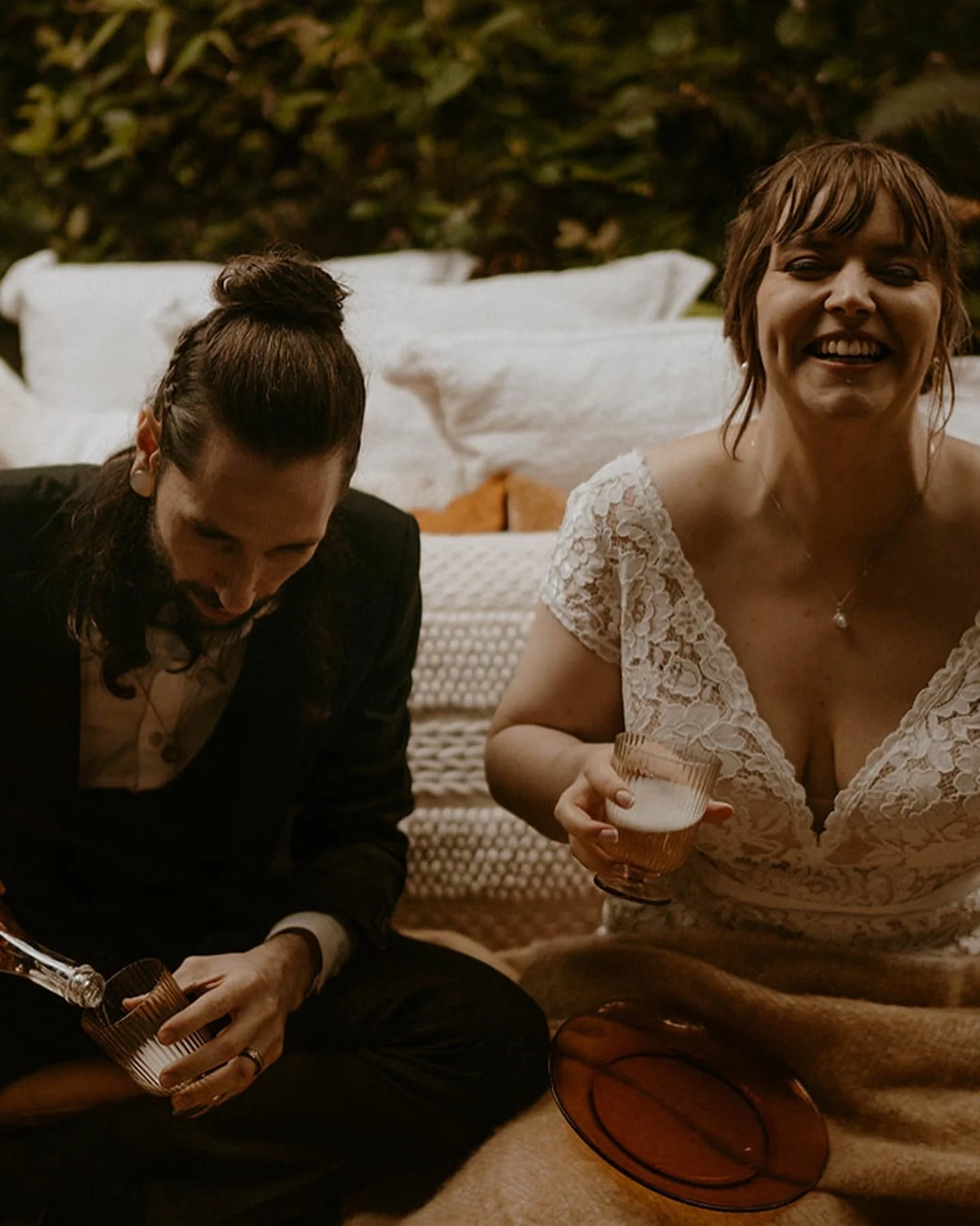 A man and a woman sitting on a blanket, laughing and holding drinks at a relaxed outdoor gathering with pillows and greenery in the background.