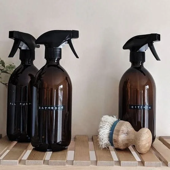 Three brown spray bottles labeled 'ALL PURPOSE', 'BATHROOM', and 'KITCHEN', with a wooden scrub brush resting against the 'KITCHEN' bottle, placed on a wooden slatted surface against a plain wall.