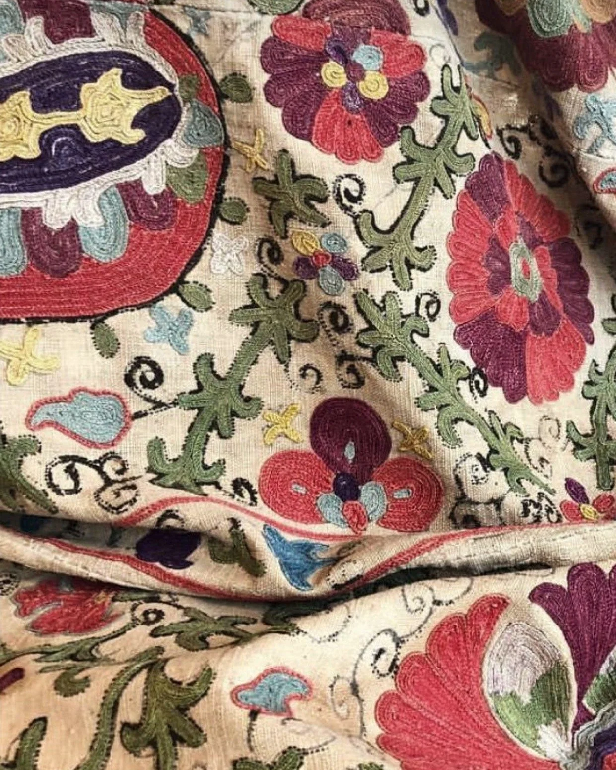 Close-up of embroidered fabric with floral and decorative patterns in red, purple, green, yellow, and blue colors on a beige background.