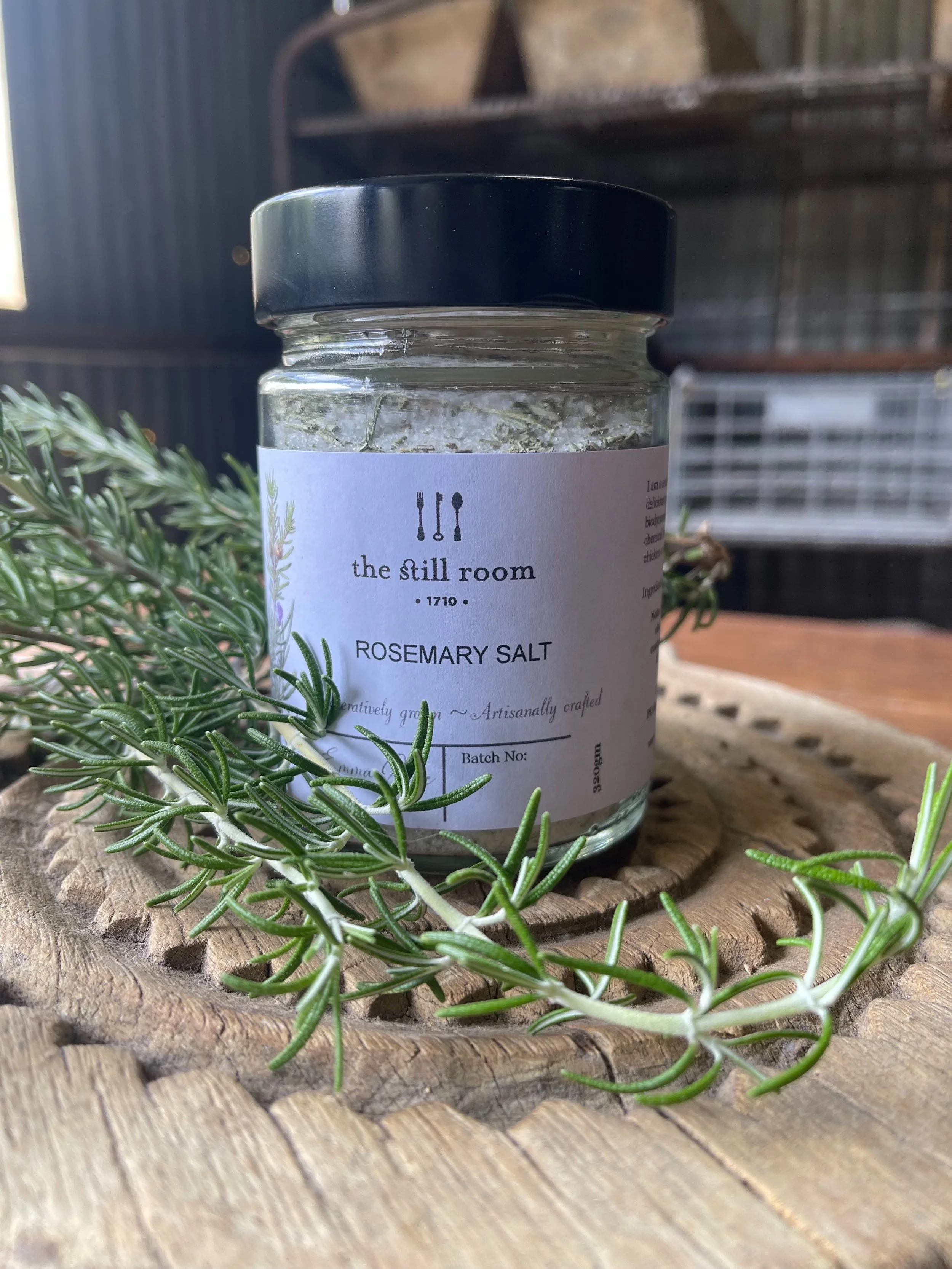 A jar of rosemary salt with a black lid, placed on a wooden surface, with green rosemary sprigs around it, and a blurred background.