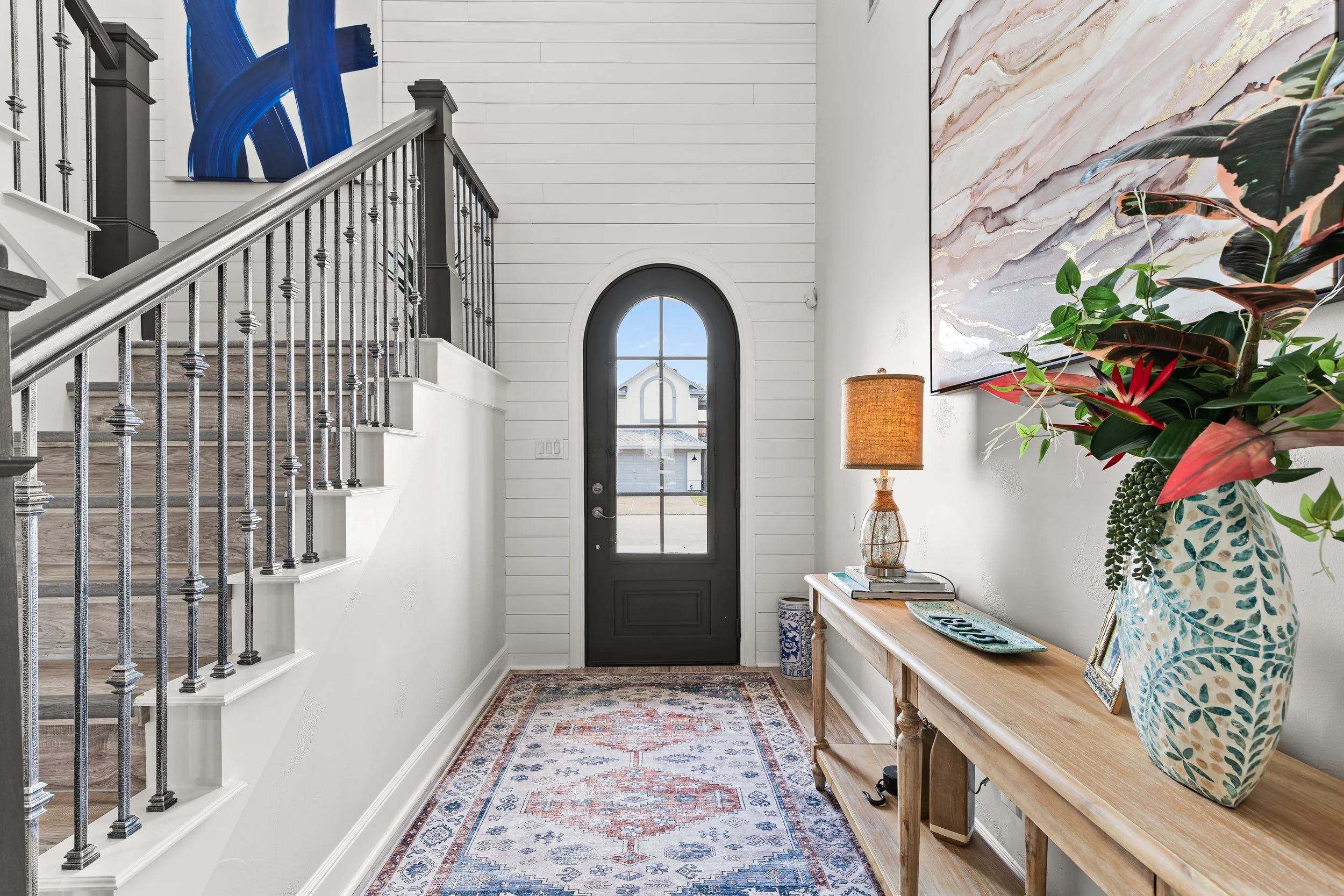 View of a modern entryway with a black front door, a wooden console table with a lamp and decorative items, a colorful indoor plant, a large abstract wall art, a patterned rug, and a staircase with black and silver railing on the left side.