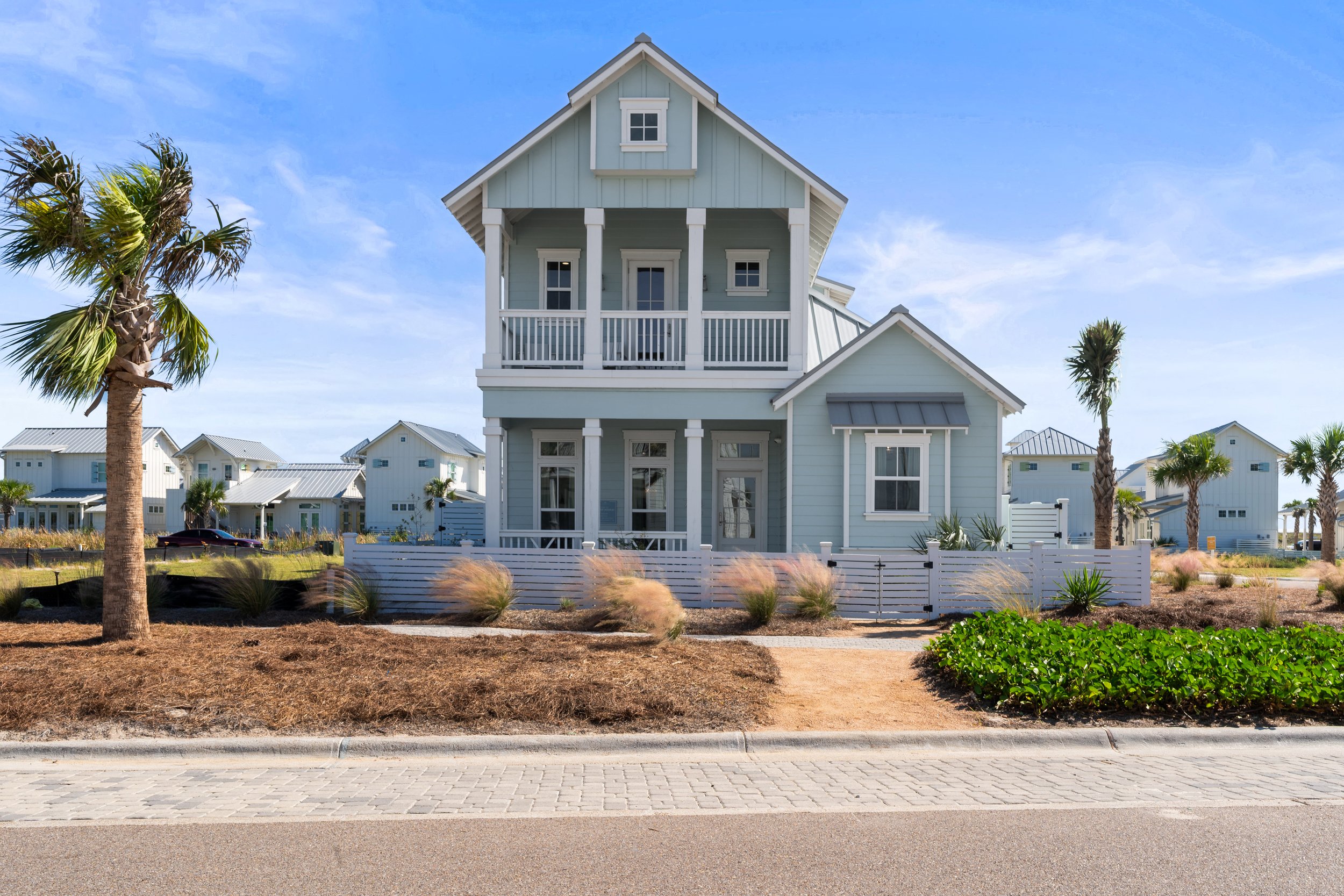 A light blue, multi-story house with white trim, a front porch, and a small balcony, set behind a white picket fence with desert landscaping, palm trees, and a brick sidewalk under a blue sky.