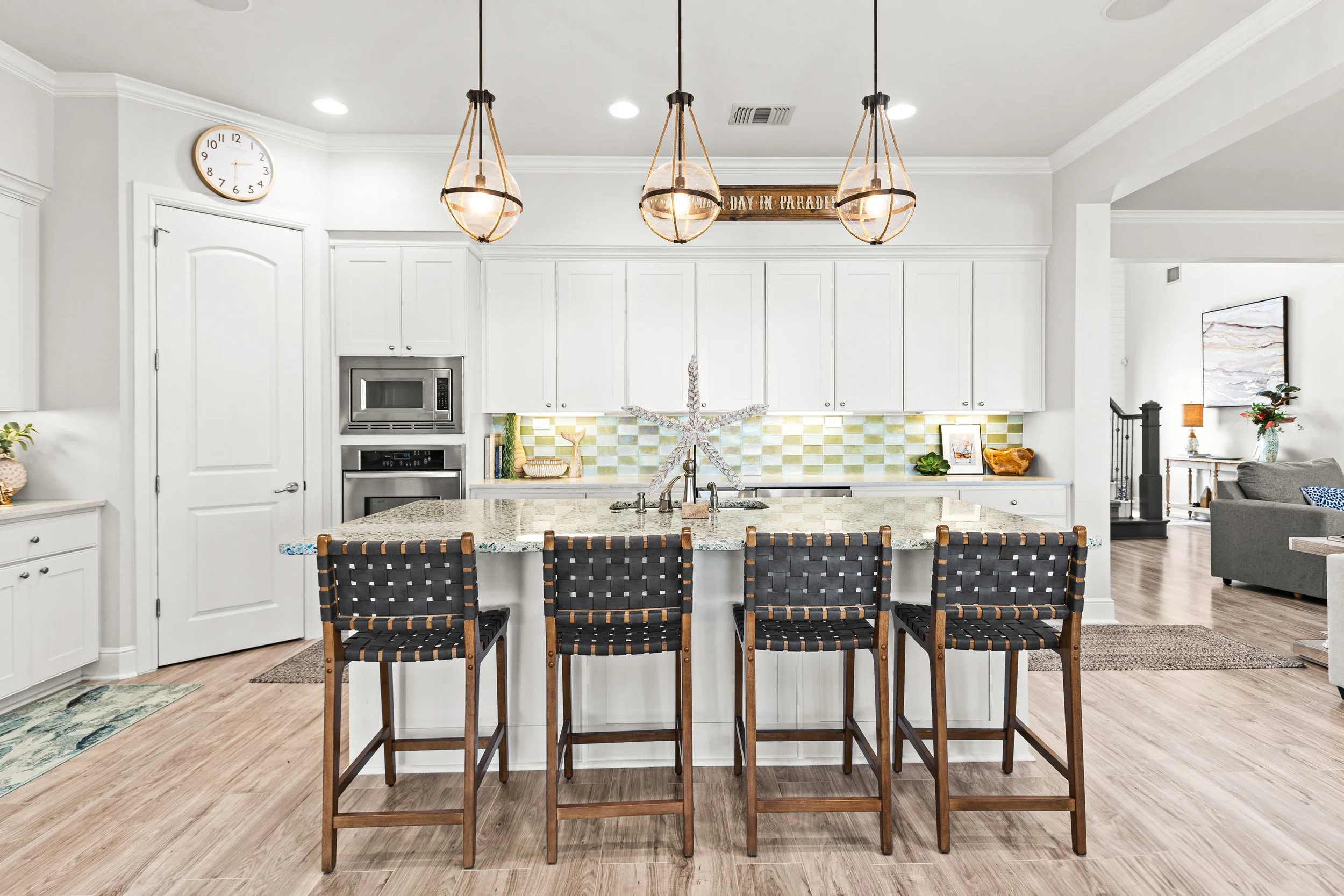 Modern kitchen with white cabinetry, granite island, pendant lights, and black woven barstools, open to a living room with gray sofa and decorative items.