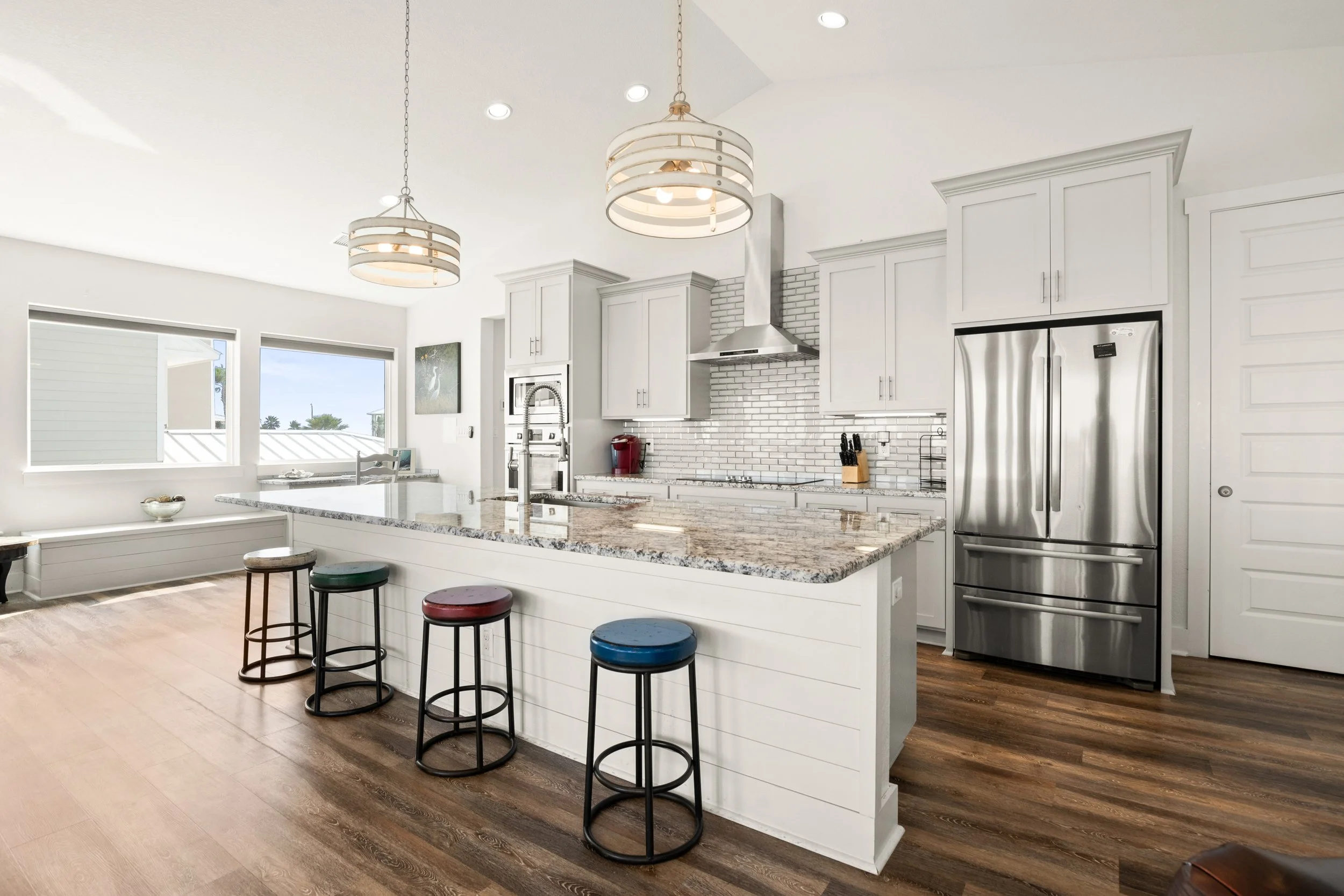 Modern white kitchen with granite countertop island, stainless steel refrigerator, and pendant lighting, with four colorful bar stools.