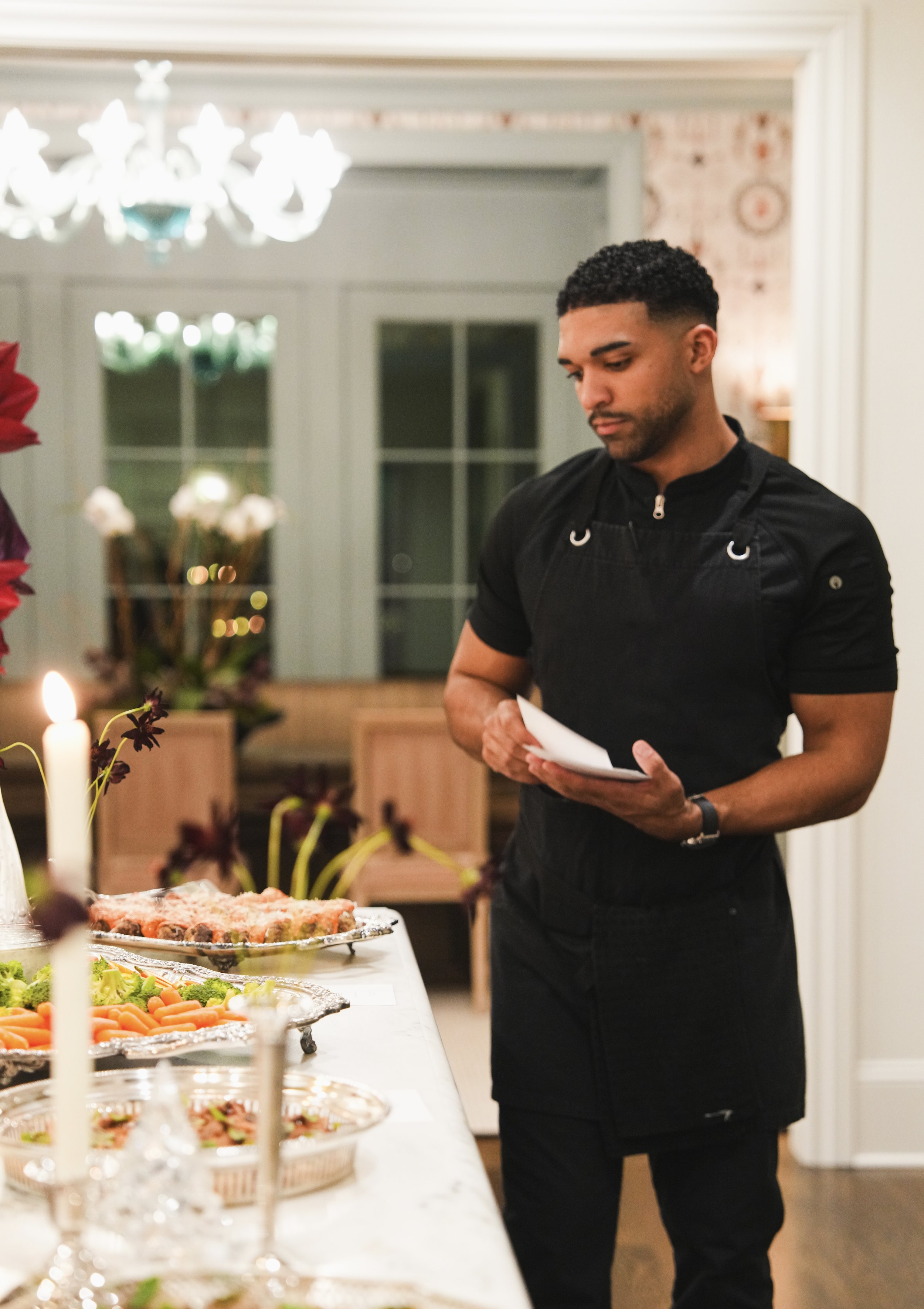 A man serving desserts in martini glasses on a wooden tray in a cozy, decorated kitchen.