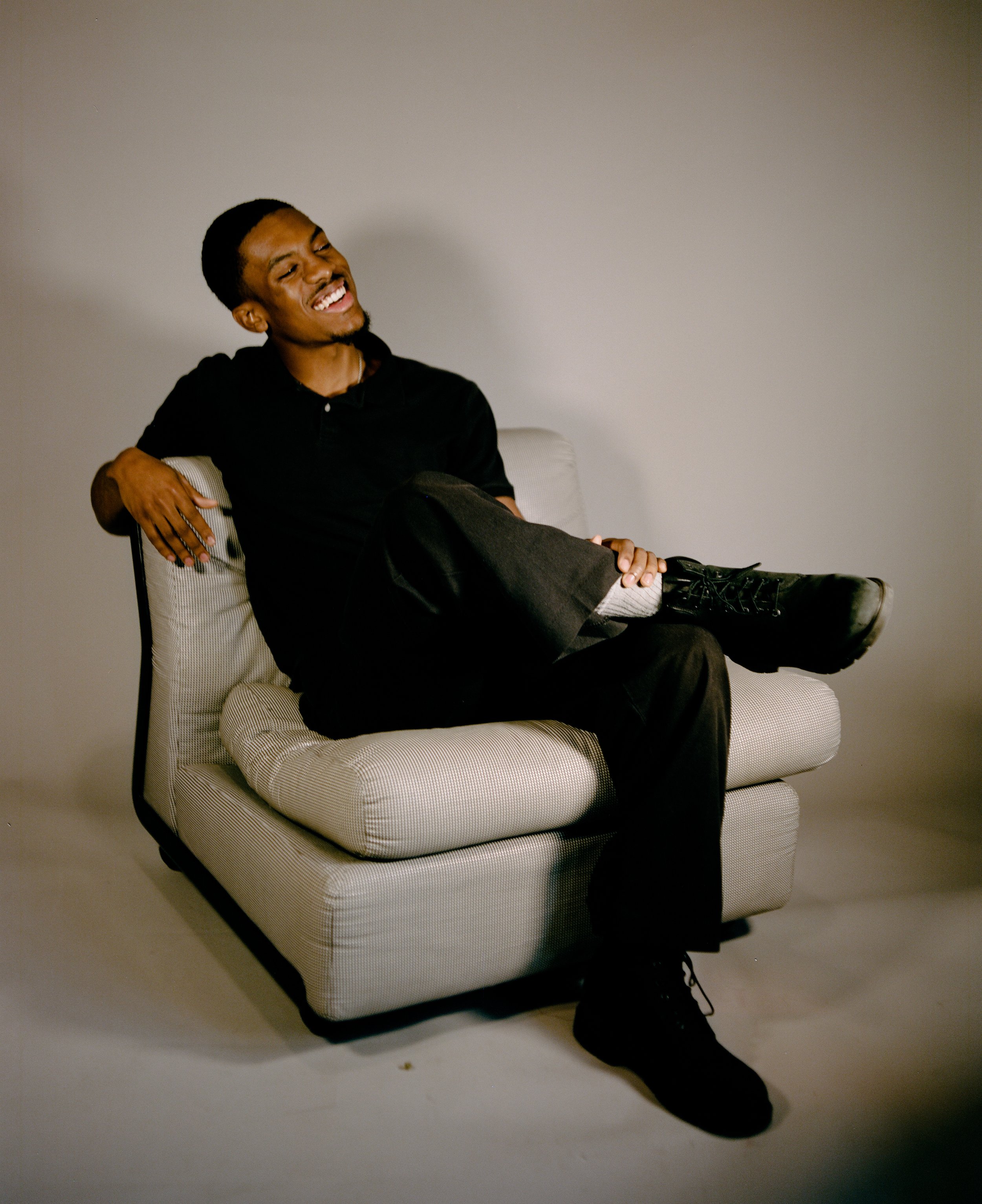 Young man with short black hair and a goatee sitting in a light-colored armchair with a striped cushion, smiling and relaxed against a plain wall background.