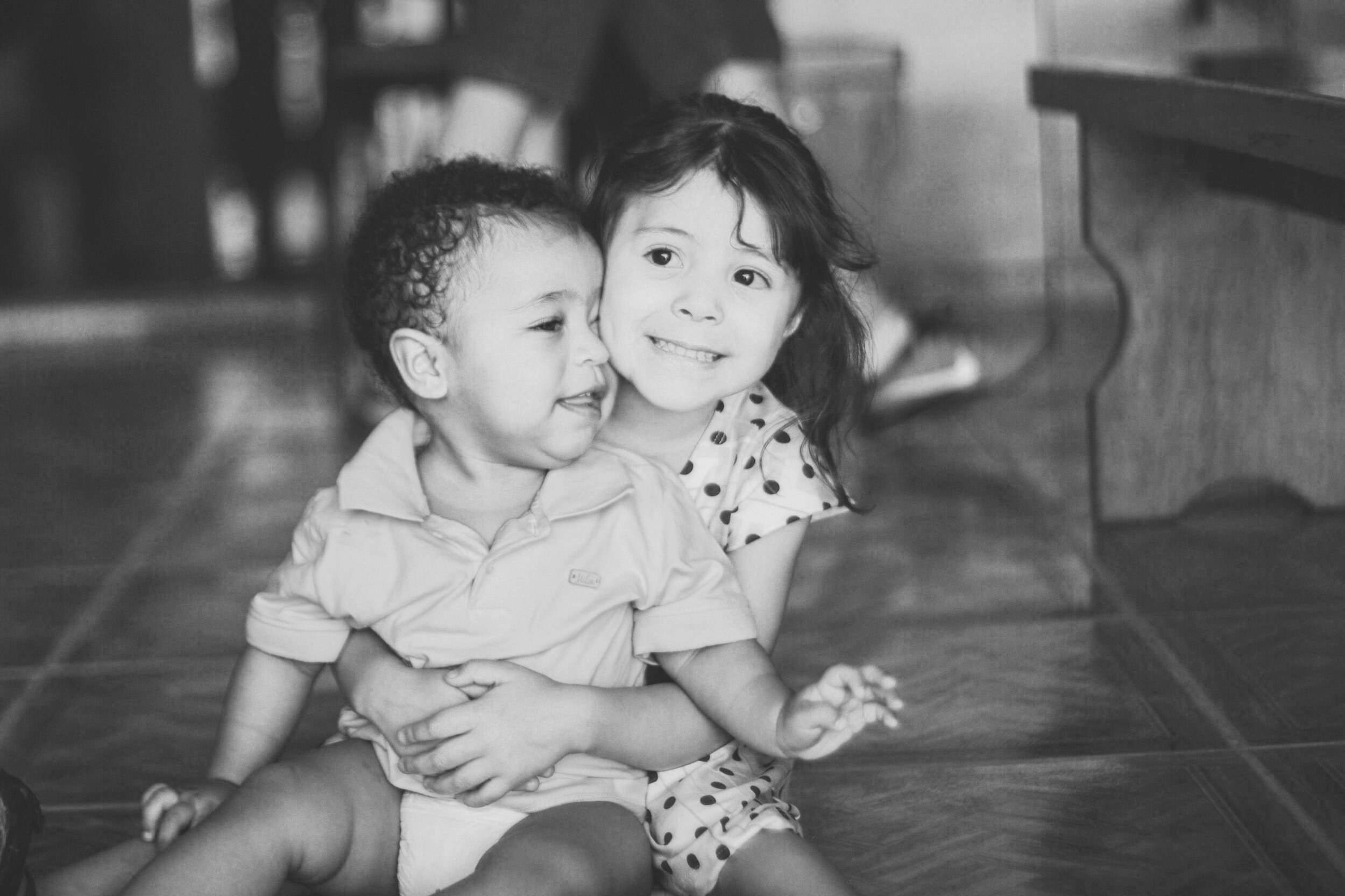 Two young children sitting on a wooden floor, smiling and hugging, with a piano in the background.