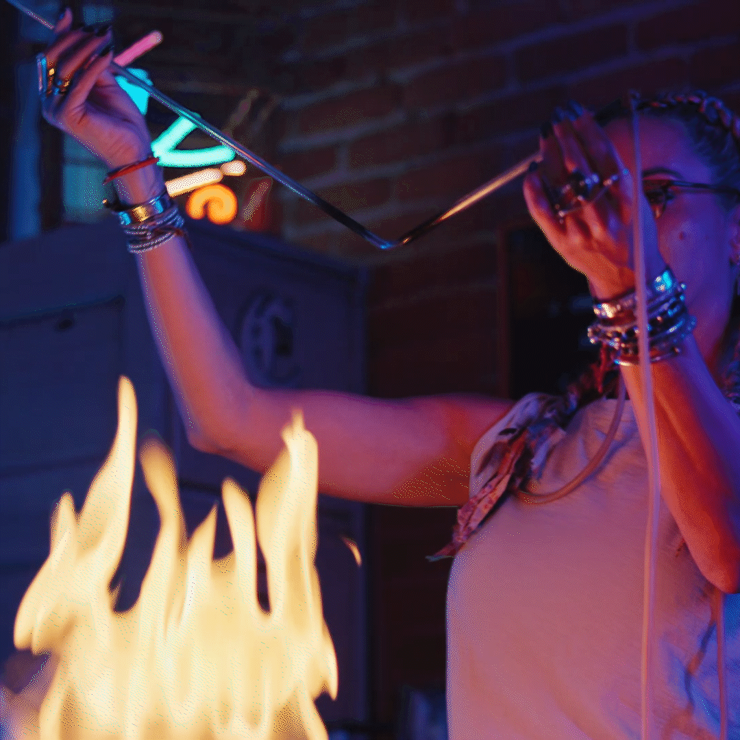A woman with braids and glasses holding a glass straw near open flames in a dimly lit environment, with brick wall in the background.