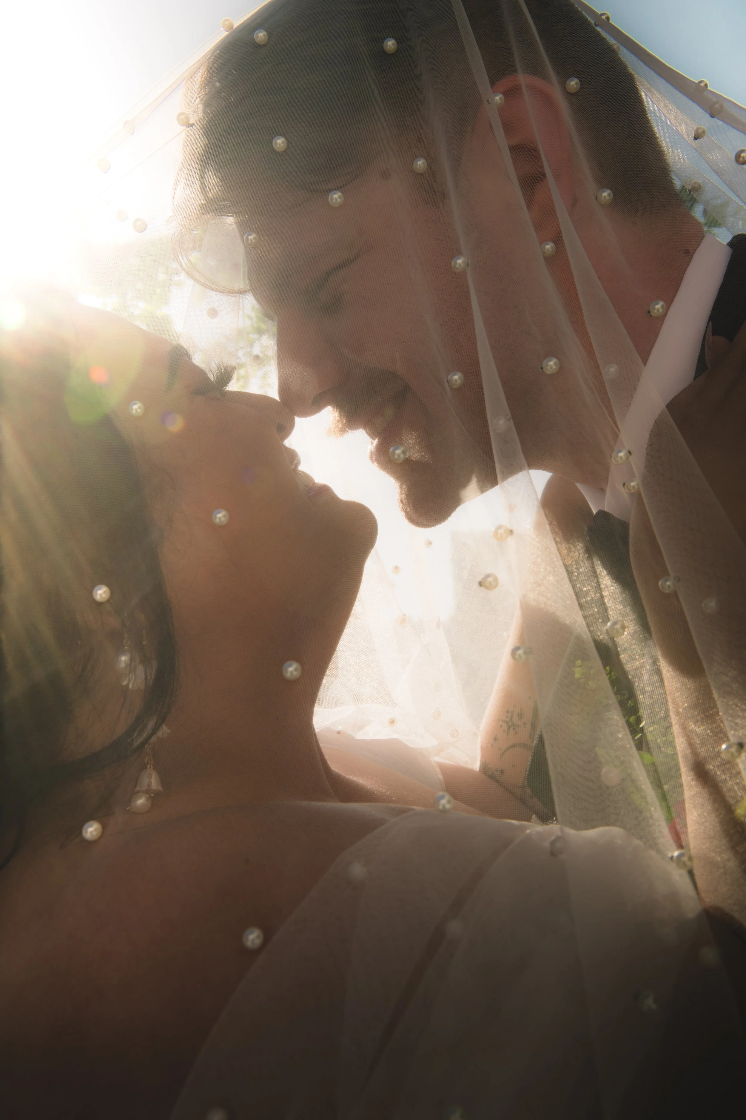 A wedding couple with their foreheads touching, smiling, under a veil with pearl beads, outdoors in sunlight. Taken in Irvine at Get Married Today.