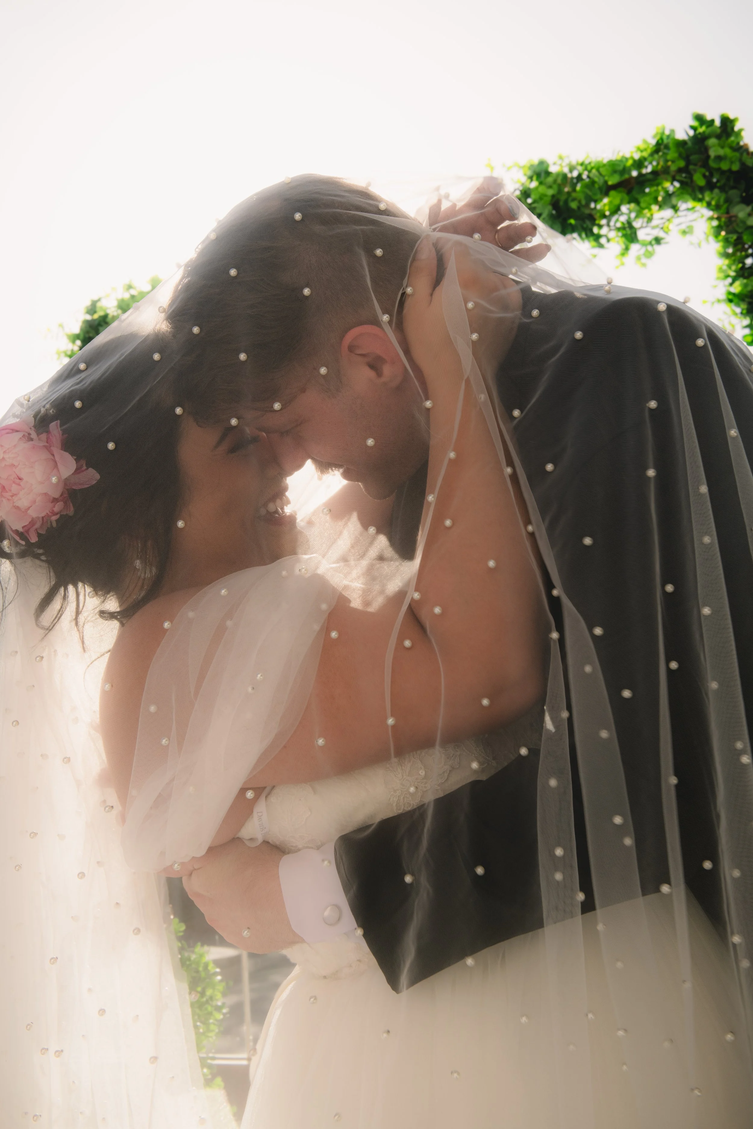 A couple, dressed for a wedding, sharing a kiss under a veil with small pearl details, outdoors with green foliage and bright sky in the background. Elopement at GetMarried.Today in Irvine, California. 