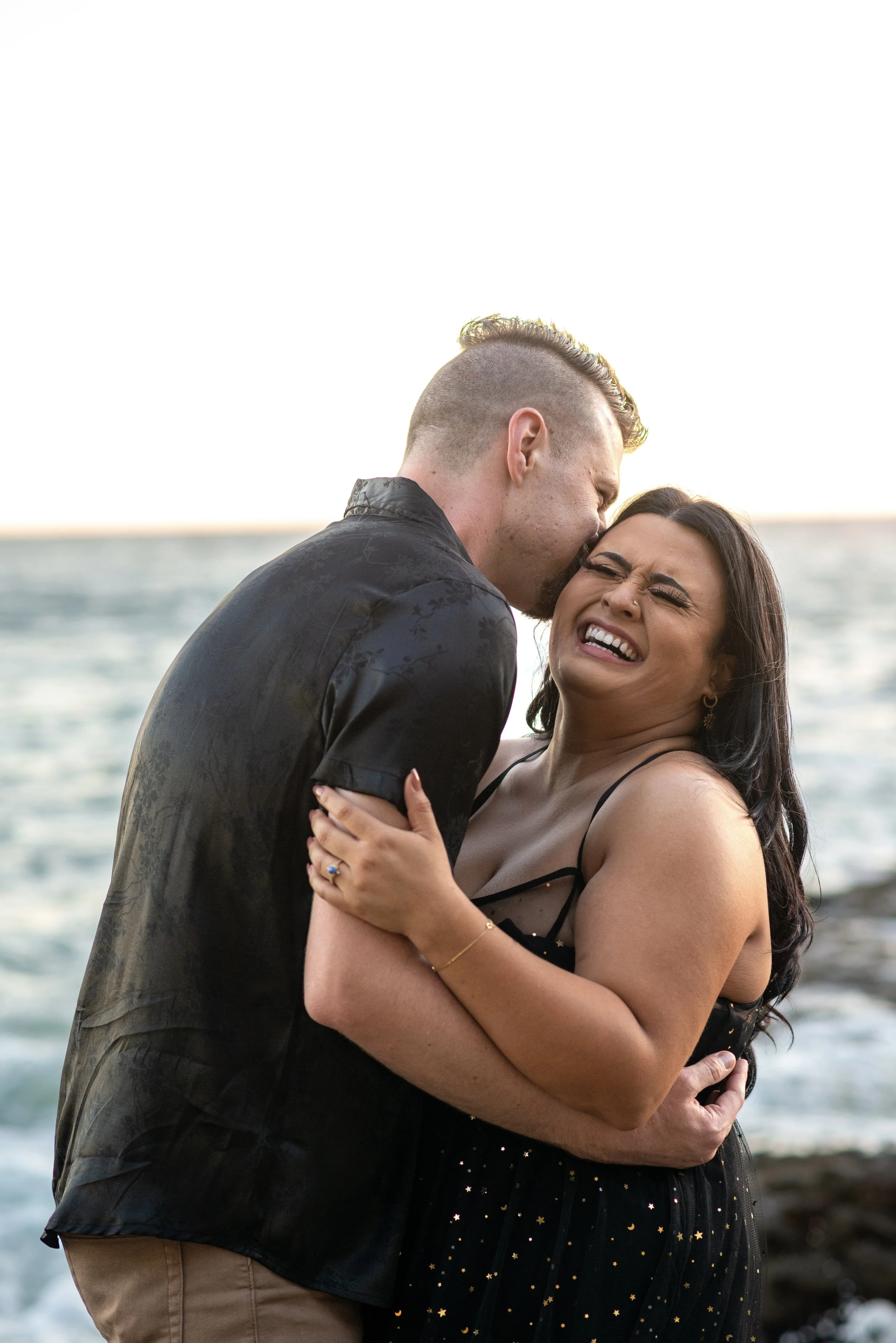 A man and a woman embrace intimately by the ocean, with the man gently kissing the woman's forehead. Both are smiling and appear joyful, dressed elegantly for a romantic outdoor setting during sunset. Shark’s cover in Laguna Beach.