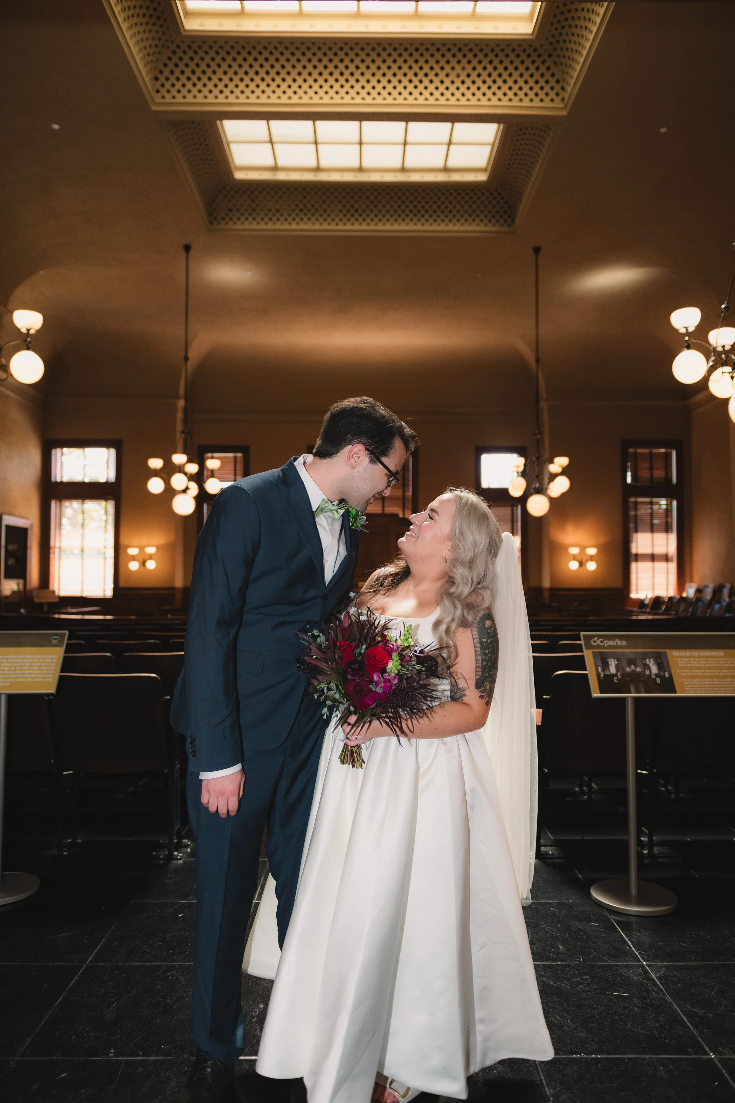 A couple stands in a church, smiling at each other. The man wears a navy suit with a light green bow tie, and the woman wears a white wedding dress and holds a bouquet of red and purple flowers. The church has tall windows and warm lighting.