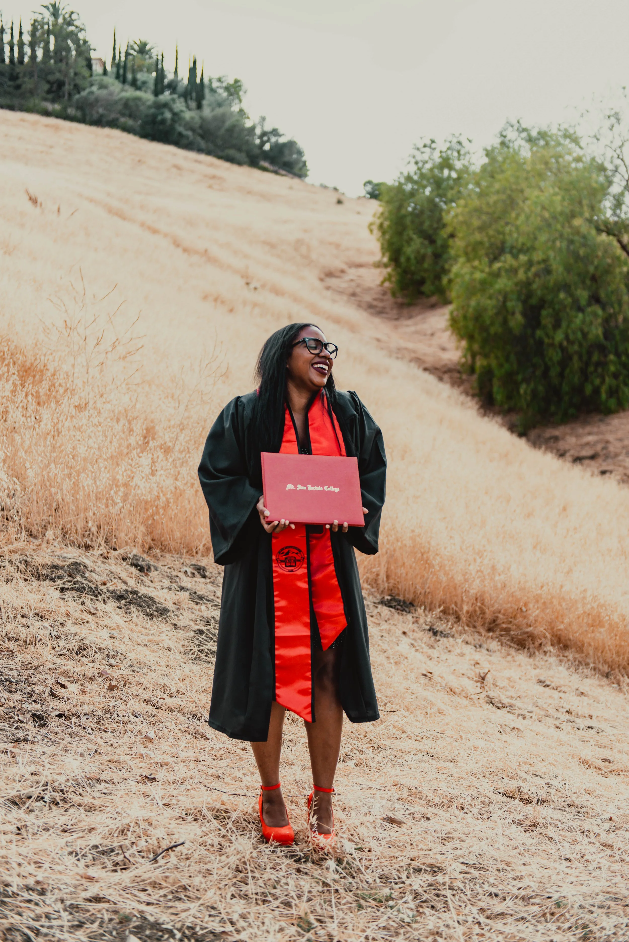 A woman dressed in a graduation gown and cap, holding a diploma, standing in a field of dried grass with trees and a hillside in the background, smiling. Mission Viejo Graduation Photographer. 