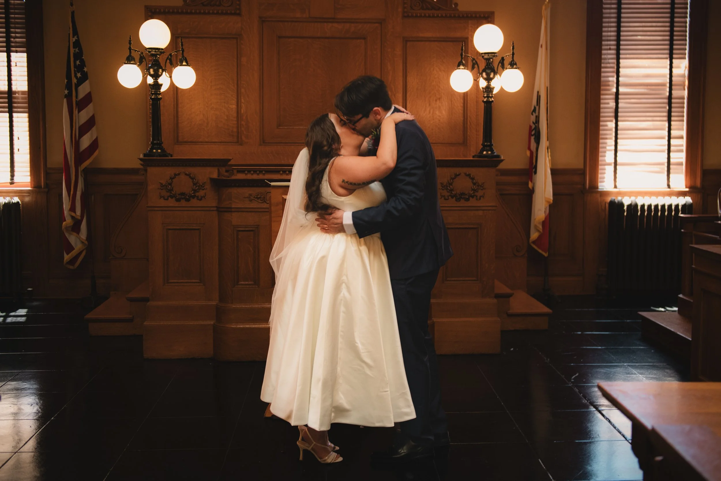 A couple is kissing in a wood-paneled room, likely during a wedding ceremony, with warm lighting and American flags visible in the background.