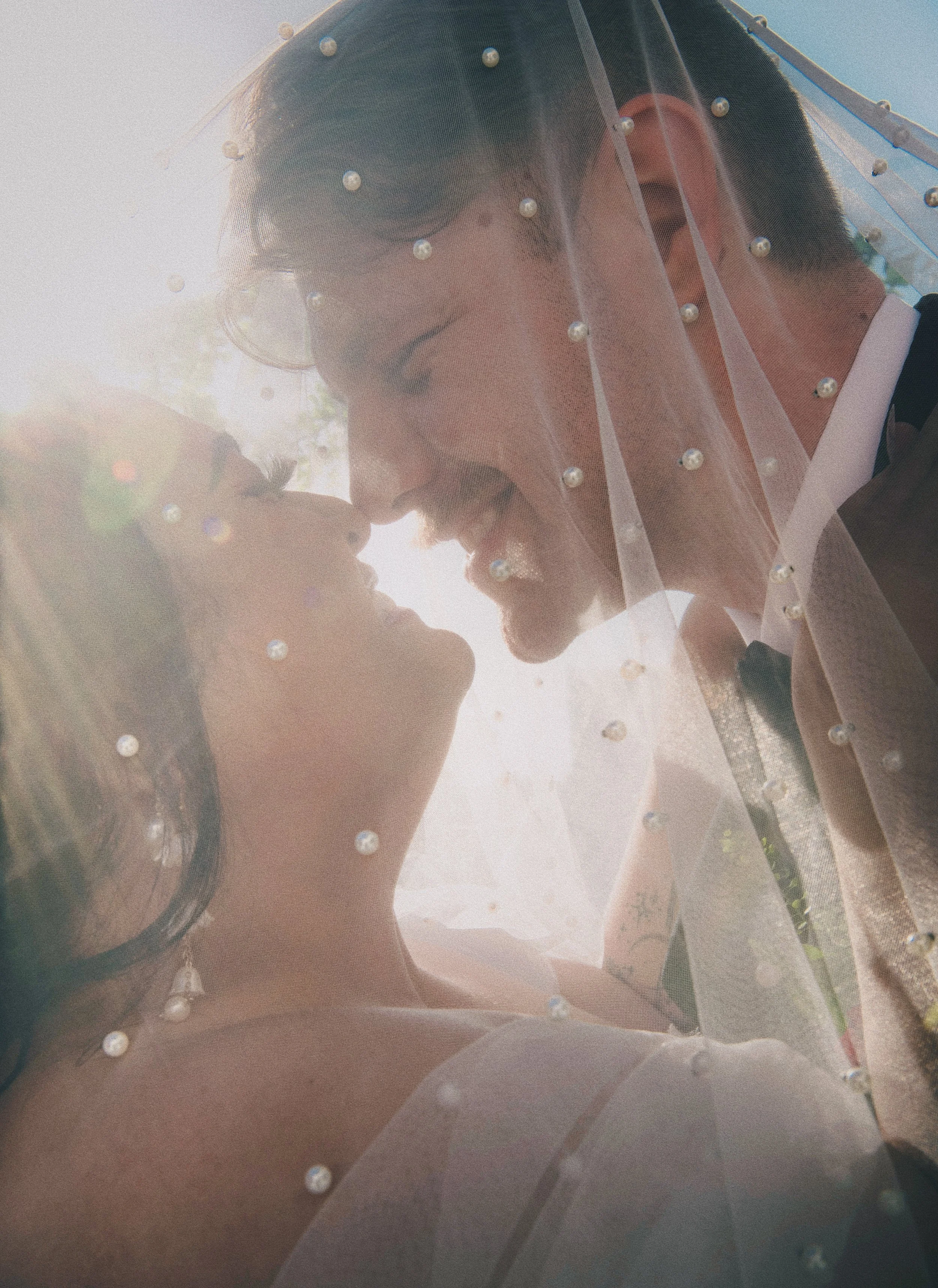 A couple with closed eyes sharing a kiss under a veil with pearl embellishments, outdoors with sunlight. Taken in Irvine, California at Get Married.Today 