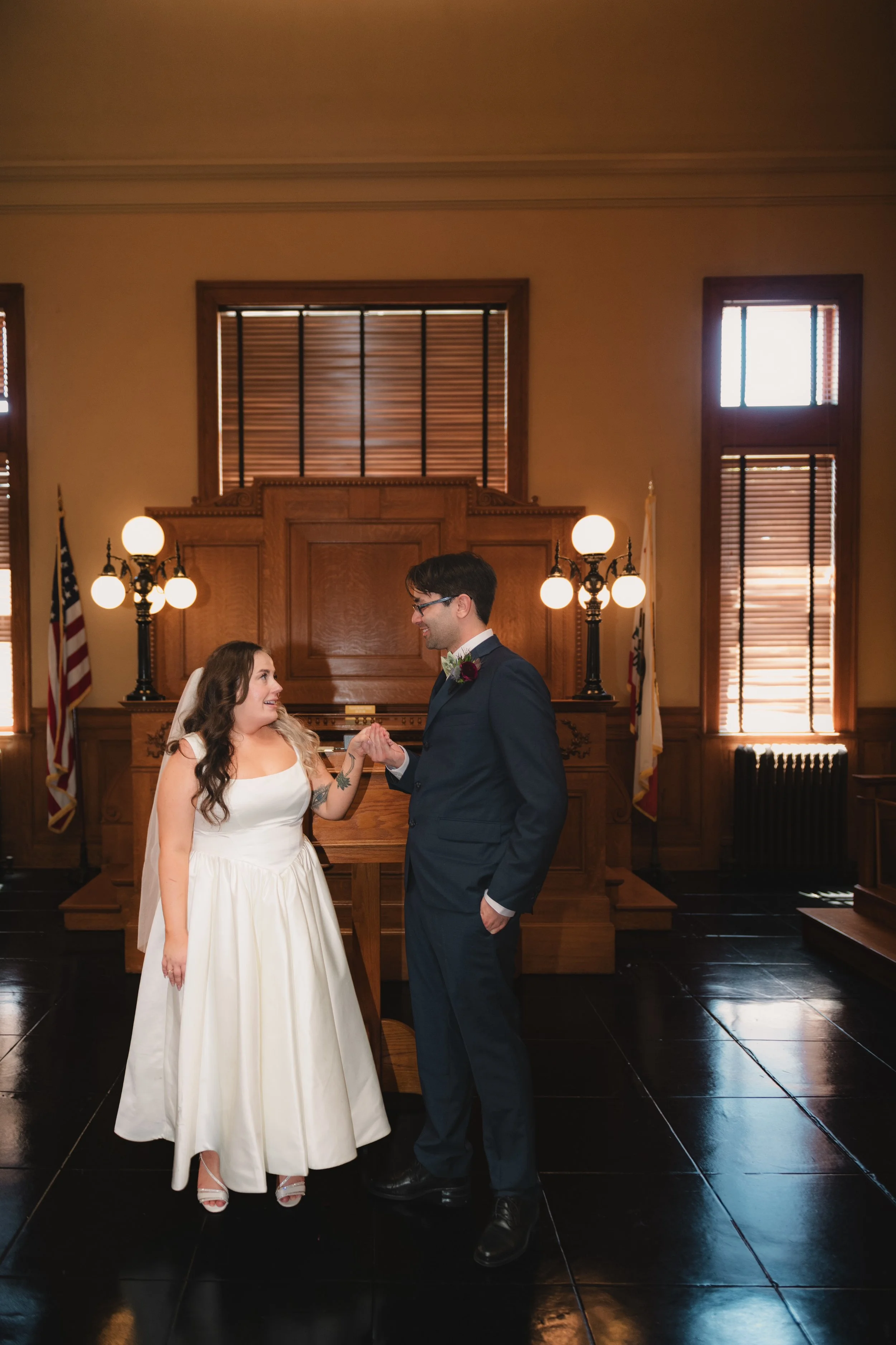 A bride and groom holding hands and facing each other inside a wood-paneled room with large windows and American flags in the background.