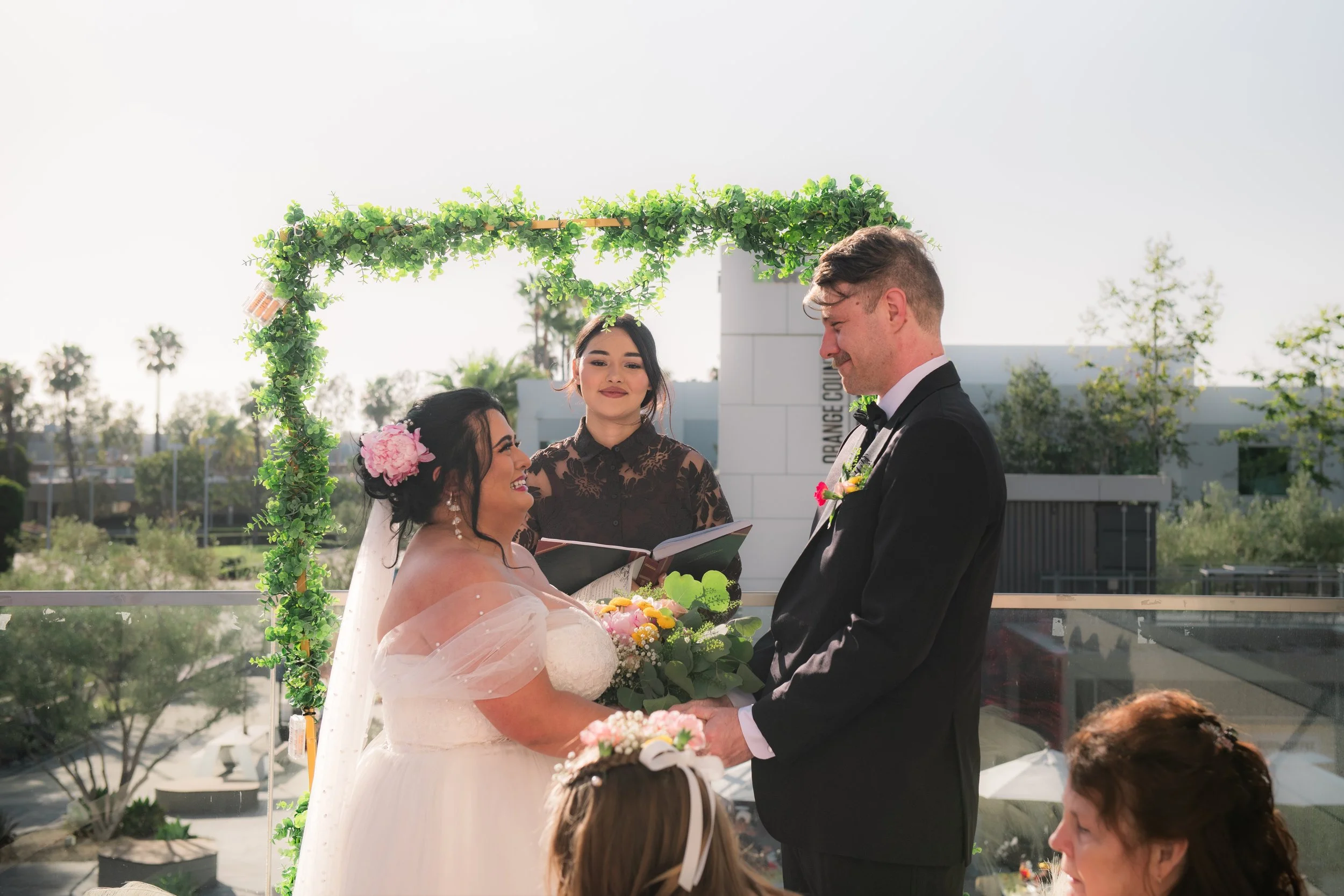 A wedding ceremony with a bride and groom exchanging vows under a green floral arch, Guests are seated and watching the couple outside with clear skies and palm trees in the background. Elopement at Get Married Today in Irvine, California.