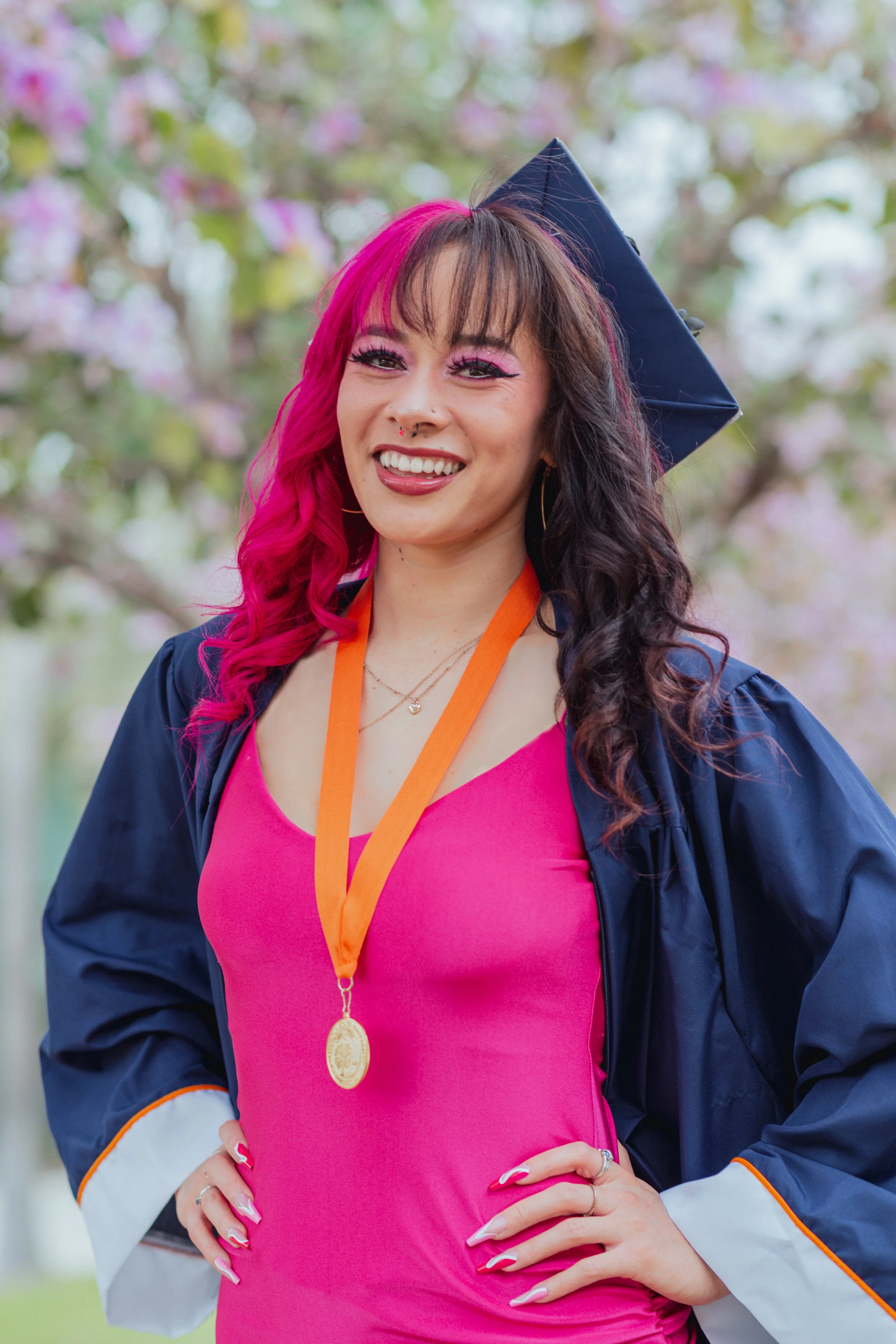 A woman in a pink dress and a dark blue graduation gown, wearing a gold medal on an orange ribbon, stands outdoors with her hand on her hip, smiling and looking at the camera, with blooming trees in the background.
