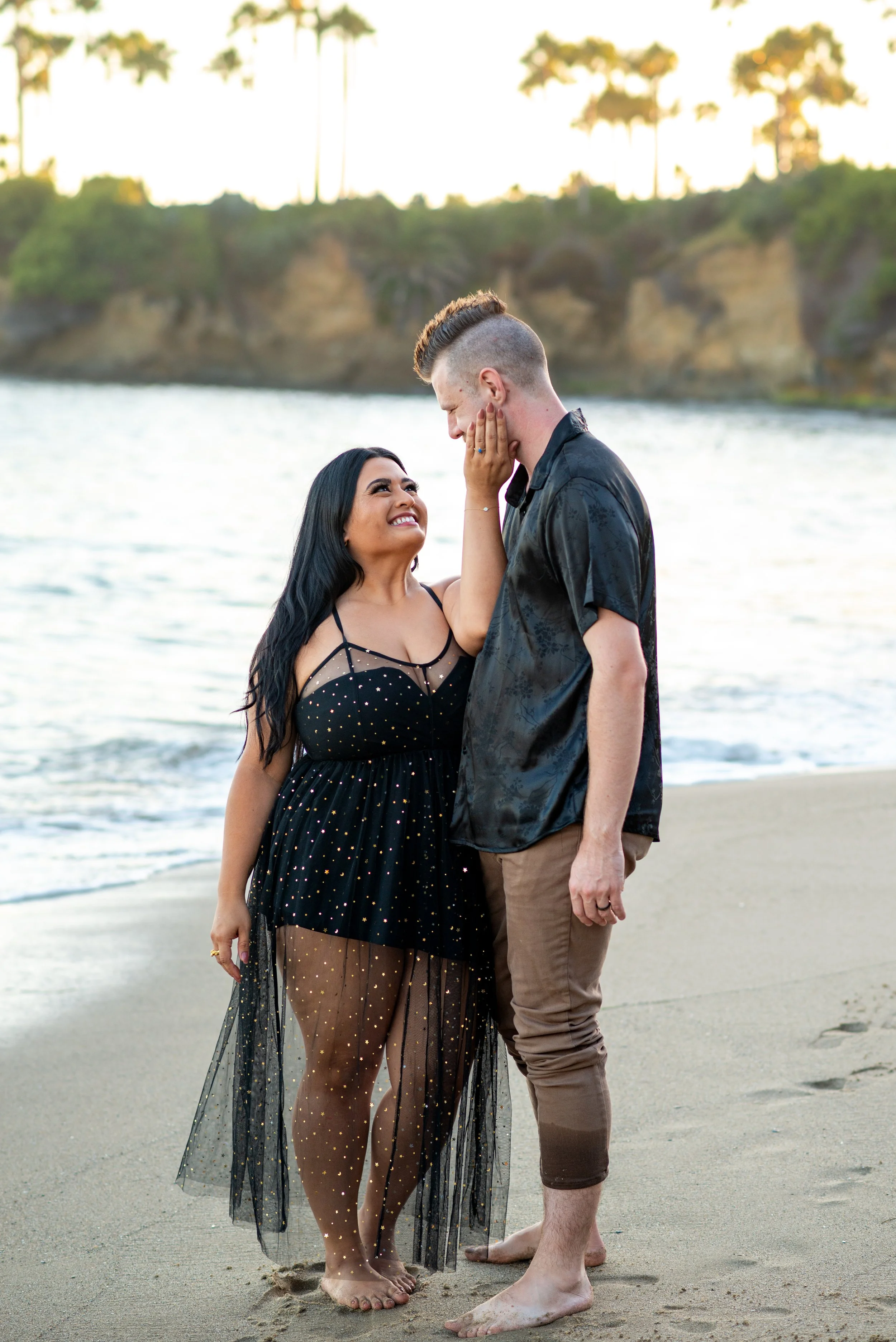 A couple standing barefoot on a beach near the water, with a woman touching the man's face and both smiling at each other, during sunset, with cliffs and palm trees in the background.