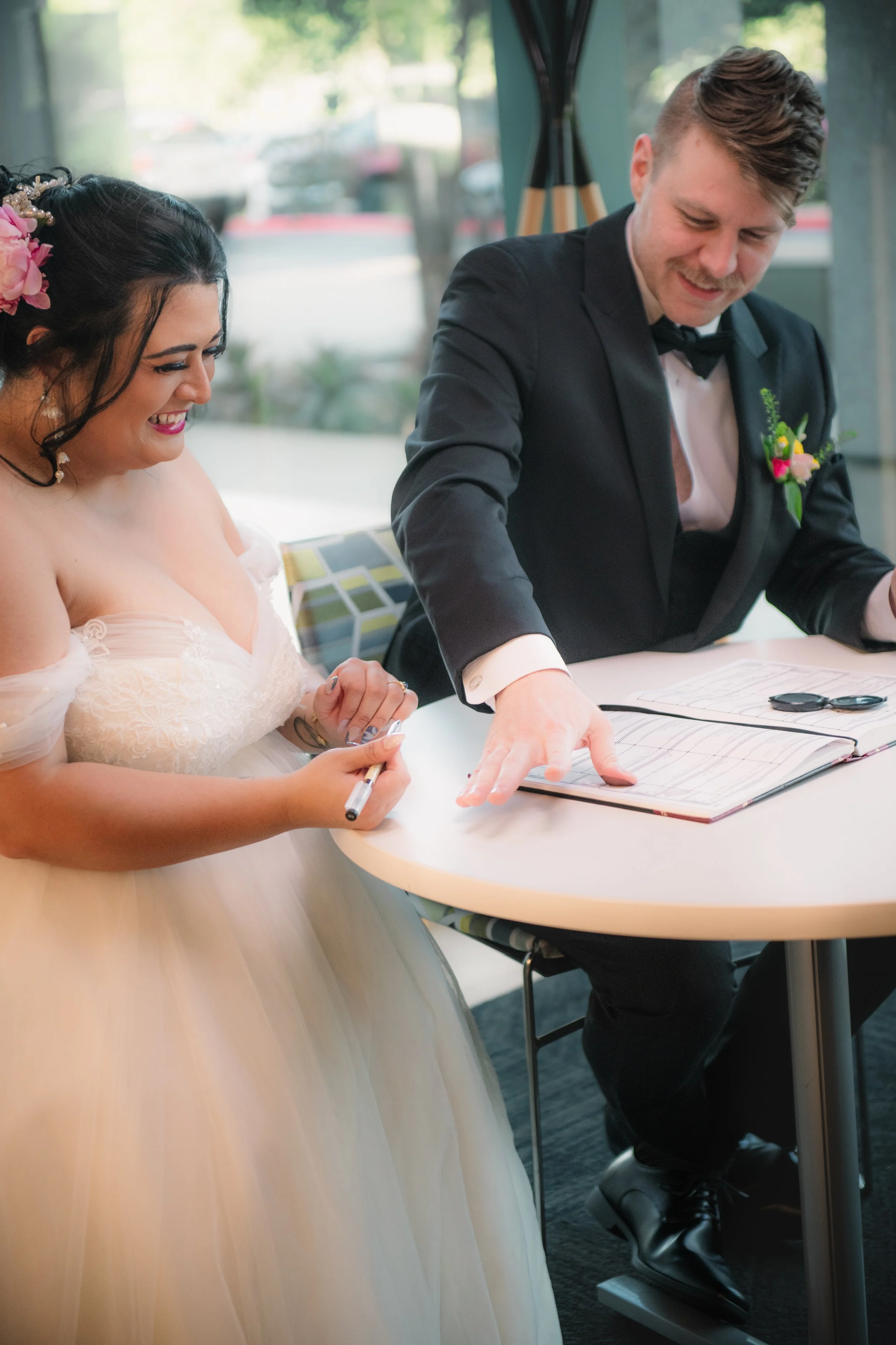 A bride and groom signing documents at a table during their wedding ceremony. The bride is smiling and wearing a white wedding dress with flowers in her hair. The groom, dressed in a black tuxedo with a boutonnière, is also smiling as he points to a 