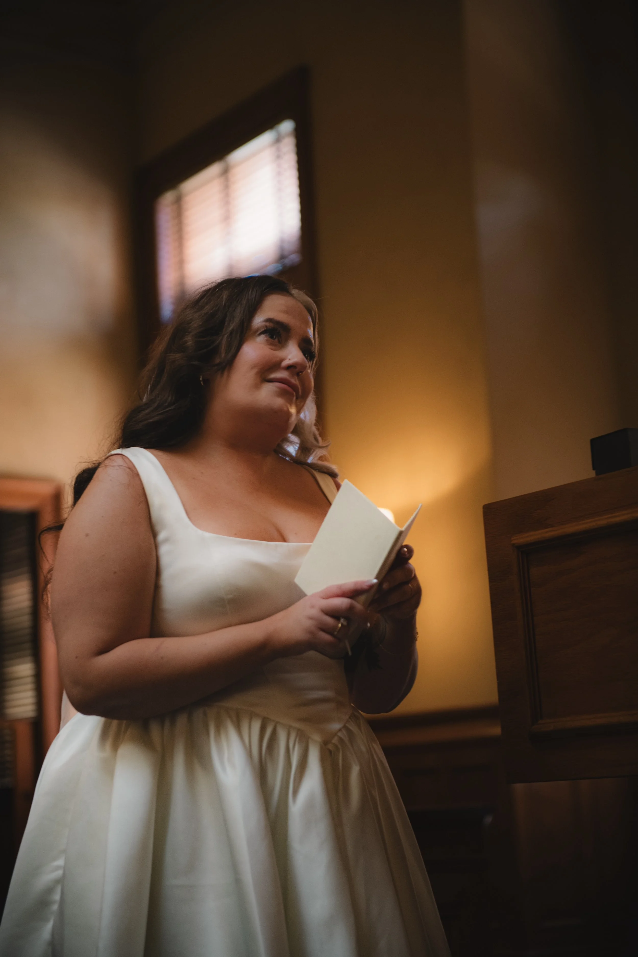 A woman in a white dress holding a piece of paper, standing indoors near a window with blinds and wooden furniture.