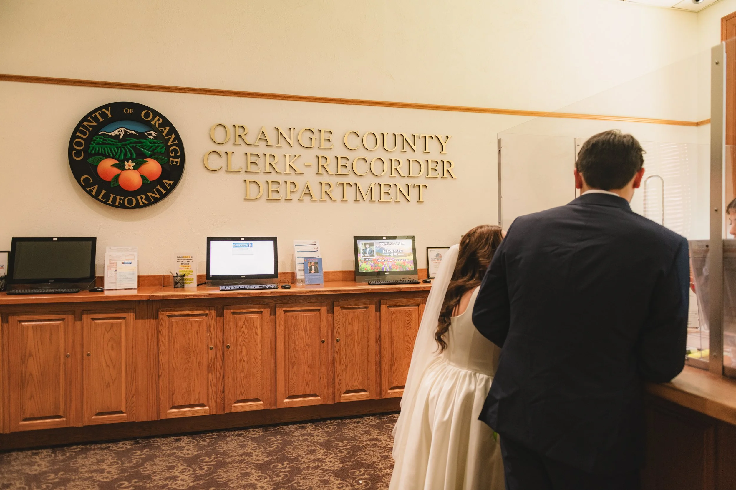 Inside the Orange County Clerk-Recorder Department with a person in a suit and a woman at the counter, computer monitors on the wooden desk, the department's logo on the wall.