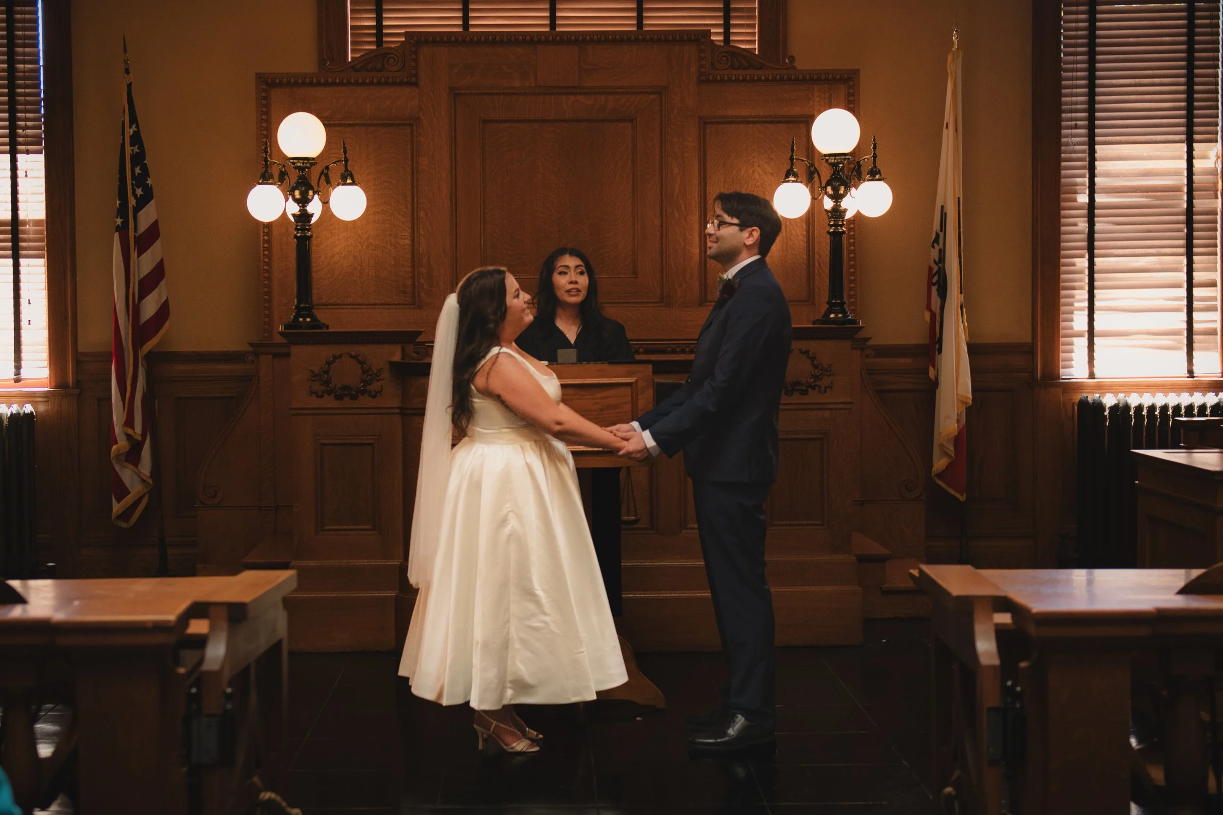 A bride and groom holding hands and looking at each other during a wedding ceremony inside Old Orange County courtroom in Santa Anna with warm lighting, and a judge or officiant standing behind them.
