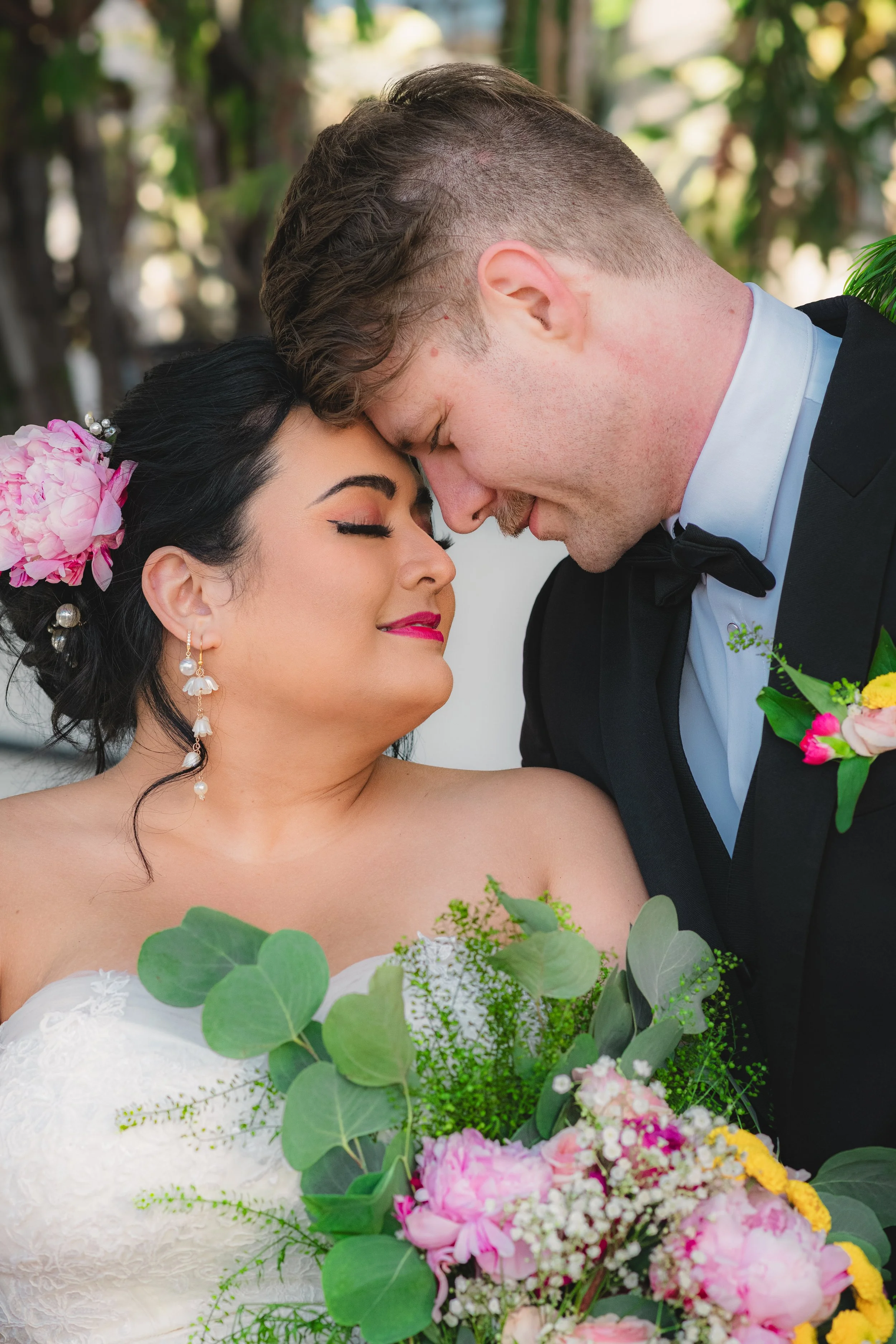 A bride and groom share an intimate moment with their foreheads touching and eyes closed. The bride has dark hair styled with pink flowers and wears earrings and a white lace dress. The groom has short, styled hair, a beard, and is dressed in a black