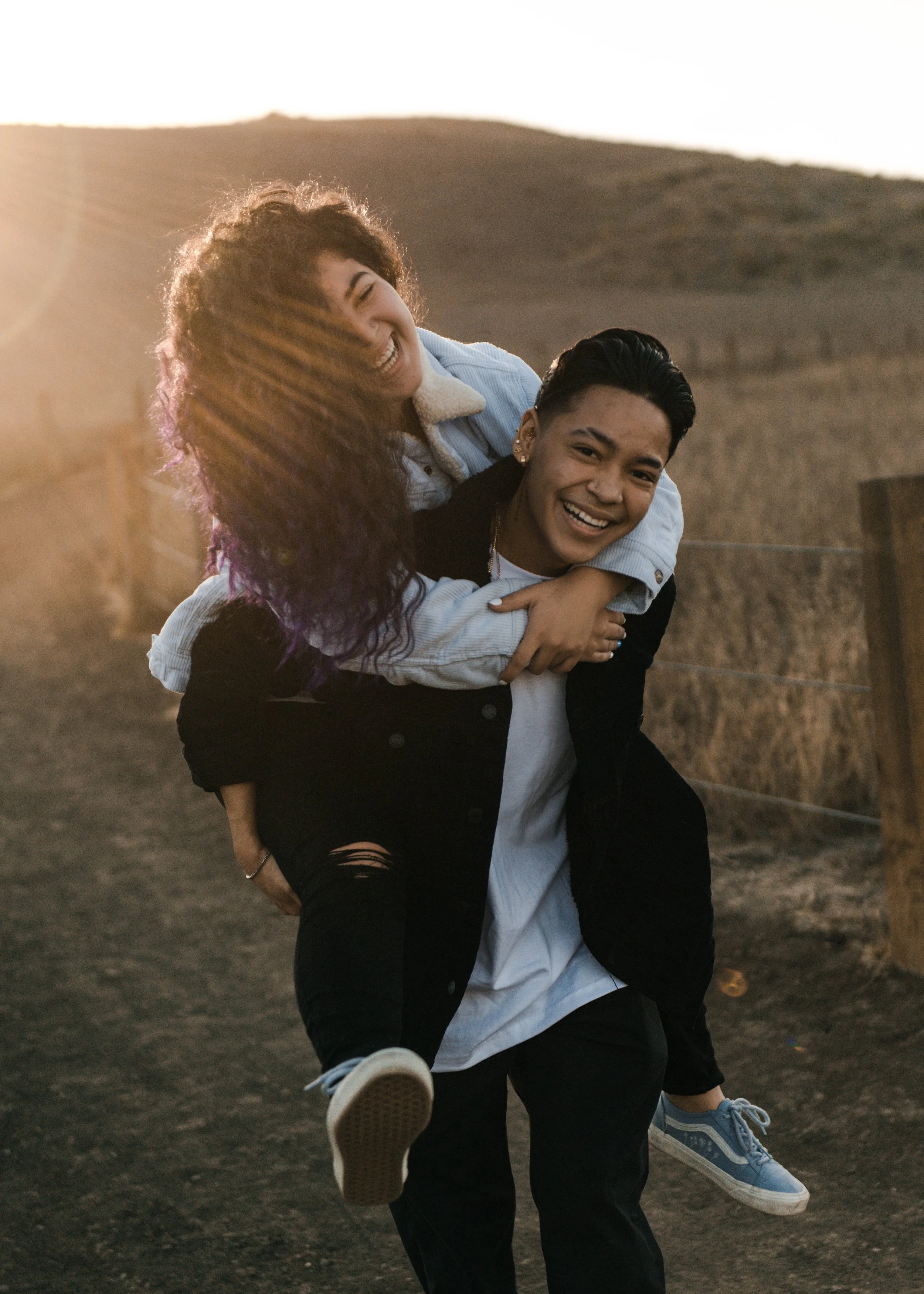 Two young women having fun in an outdoor setting during sunset, with one giving the other a piggyback ride on a dirt path with a hilly landscape in the background.