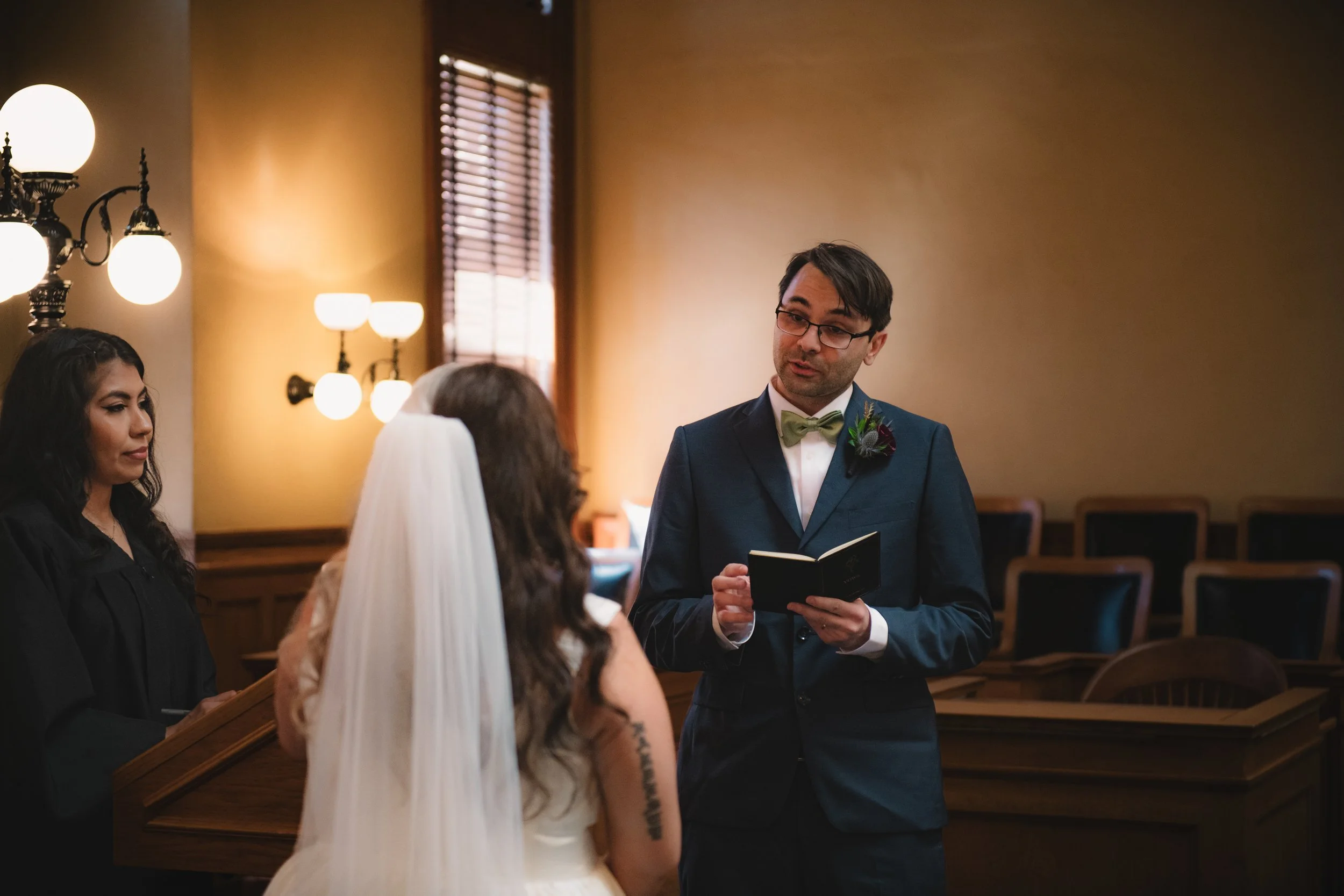 A wedding ceremony taking place in a church with an officiant reading vows to the bride and groom, who are facing each other. The bride has long hair, a veil, and a white dress, while the groom is wearing glasses, a dark suit, and a green bow tie. A 