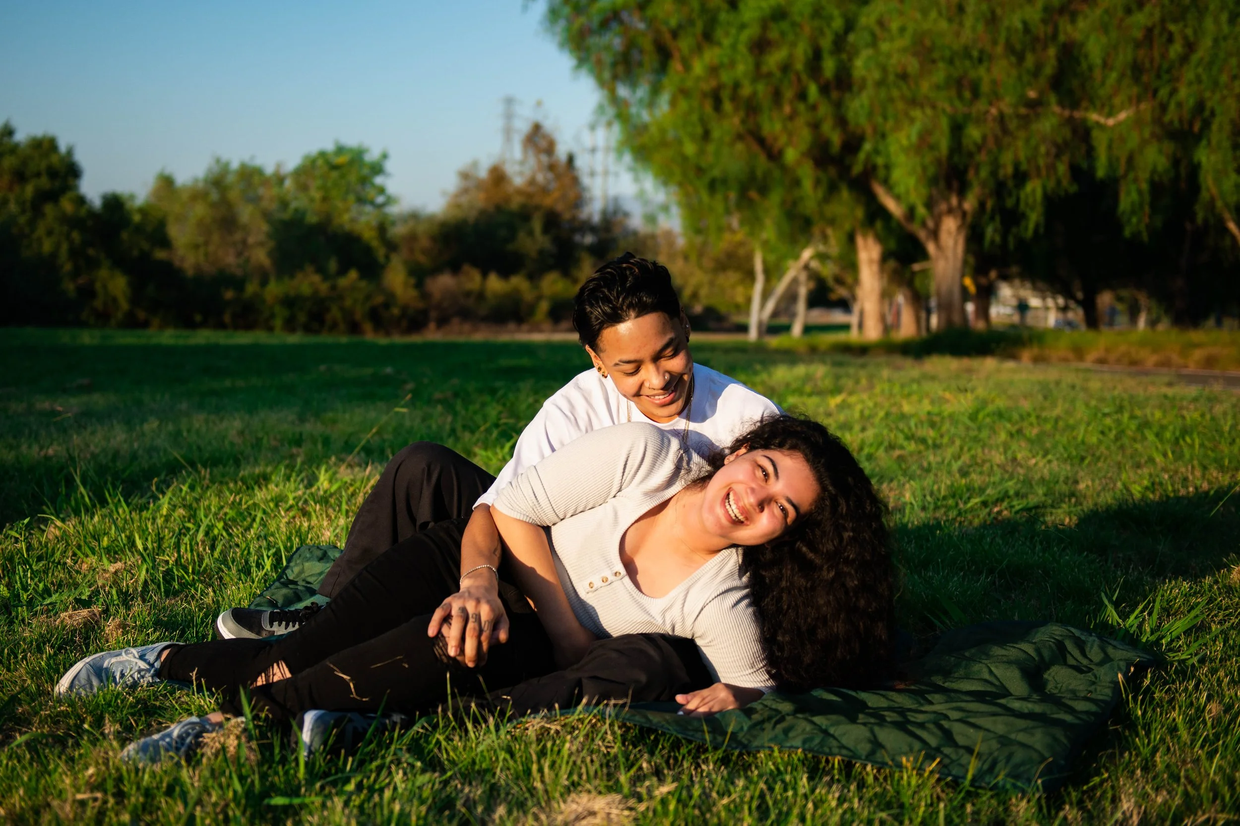 Two women lying on grass in a park, smiling and laughing, with trees and blue sky in the background.