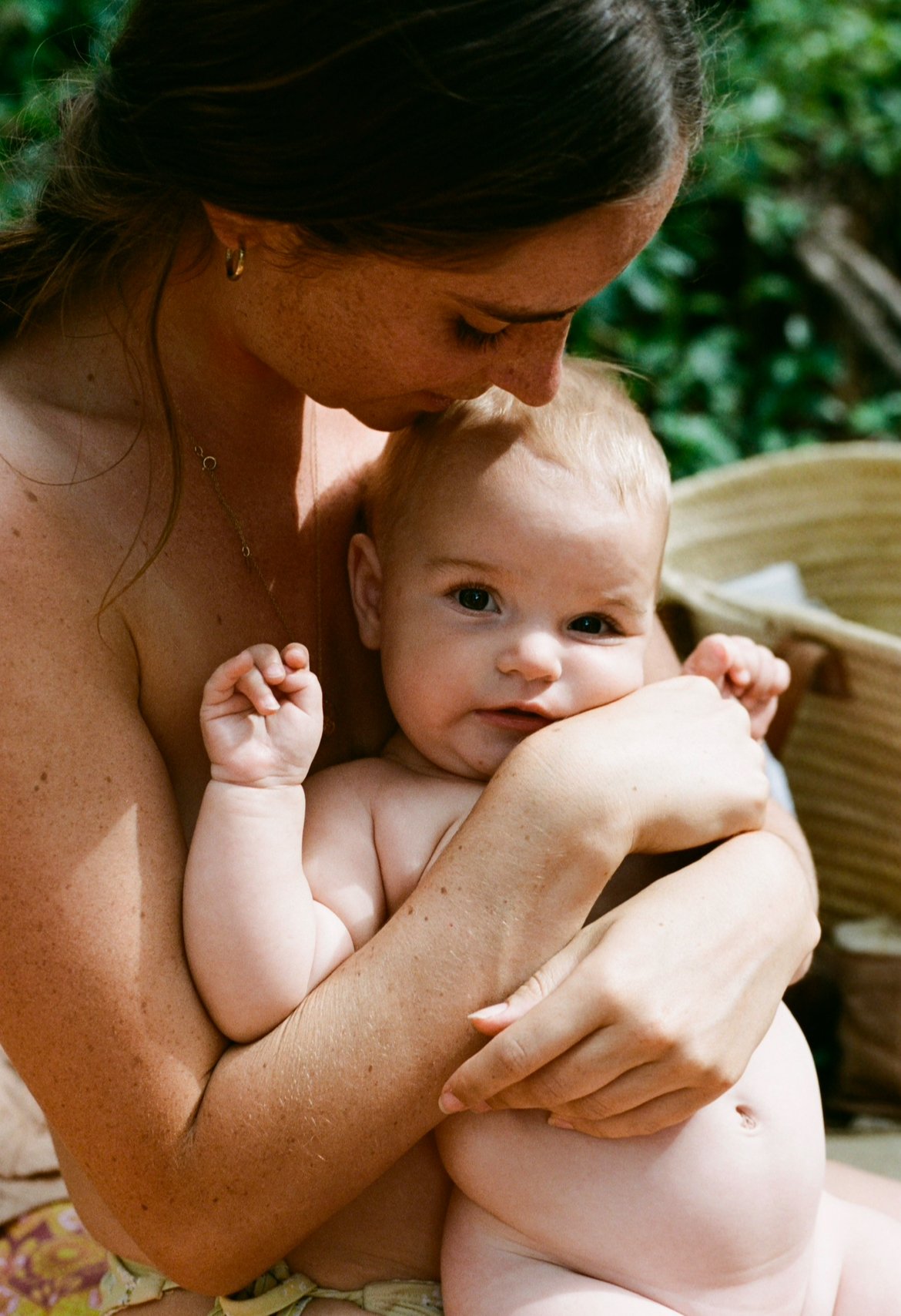 A woman with dark hair holding a naked baby with light hair, brown eyes, and a round belly. The woman is looking at the baby, who is looking at the camera, with a gentle expression. The background has green foliage and a wicker basket.