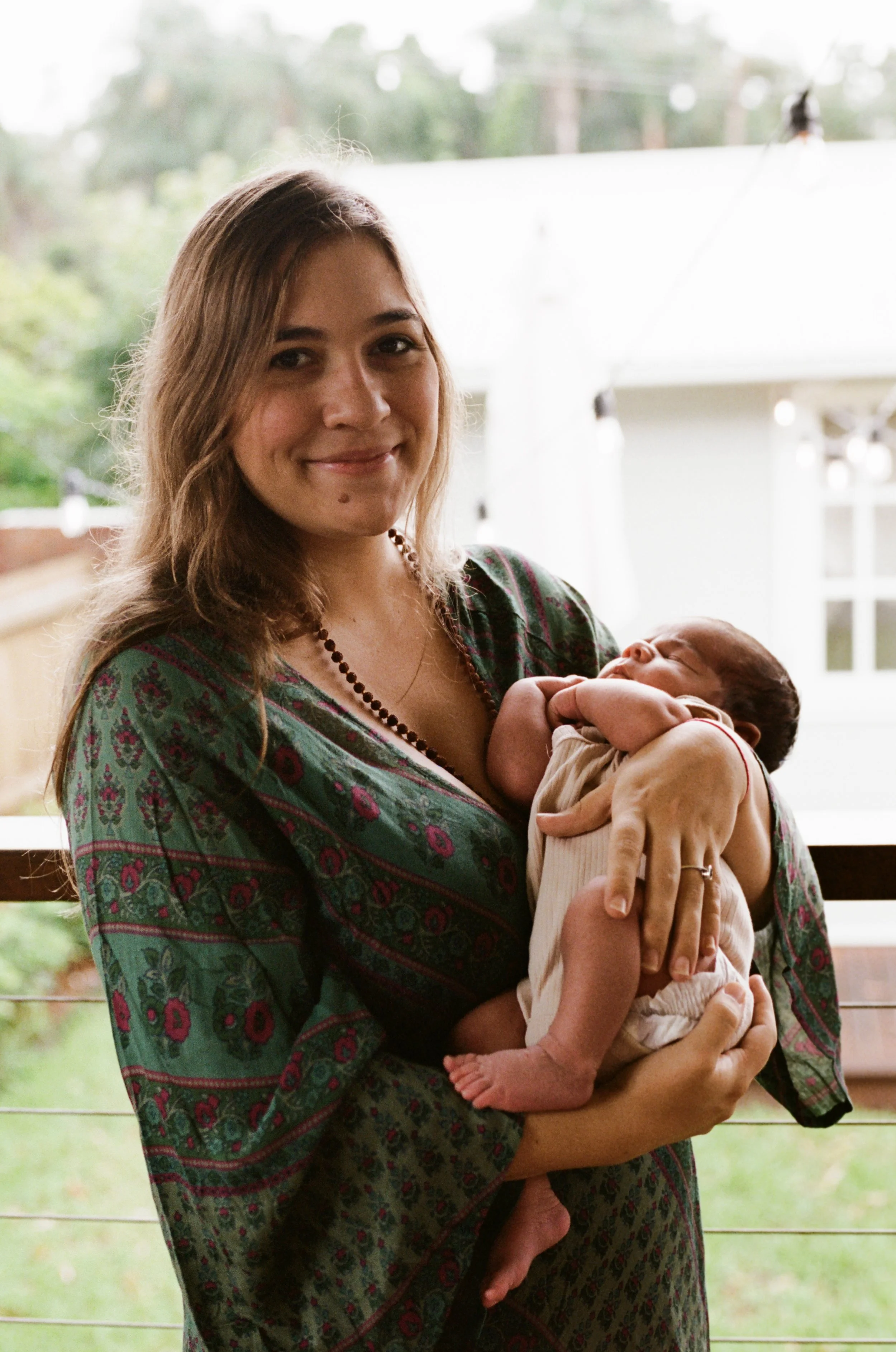 A woman with long brown hair wearing a patterned dress holds a small baby in her arms, smiling softly inside a house or porch with a blurred background of trees and a white house.