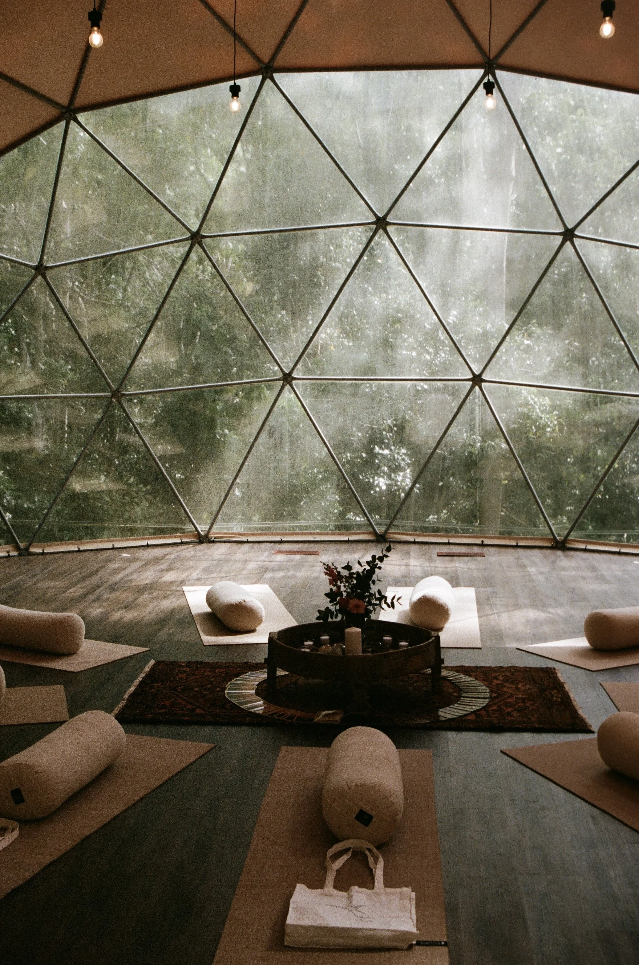 Indoor yoga or meditation studio with large geodesic dome glass ceiling showing rain outside. Yoga mats, bolsters, a blanket, and a small table with flowers and candles are arranged for a meditation or yoga session.