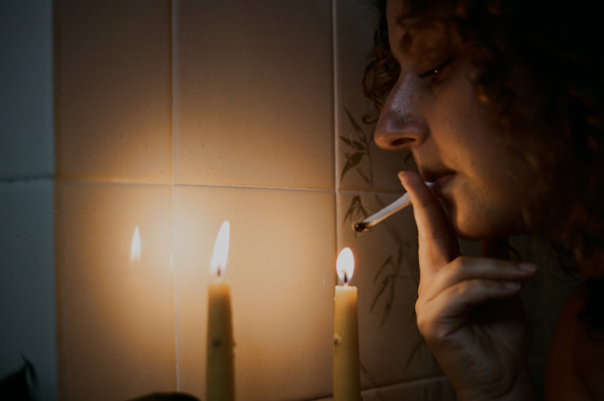 A person with curly hair and earrings is lighting a candle with a match, with other lit candles beside them, against a tiled wall background.