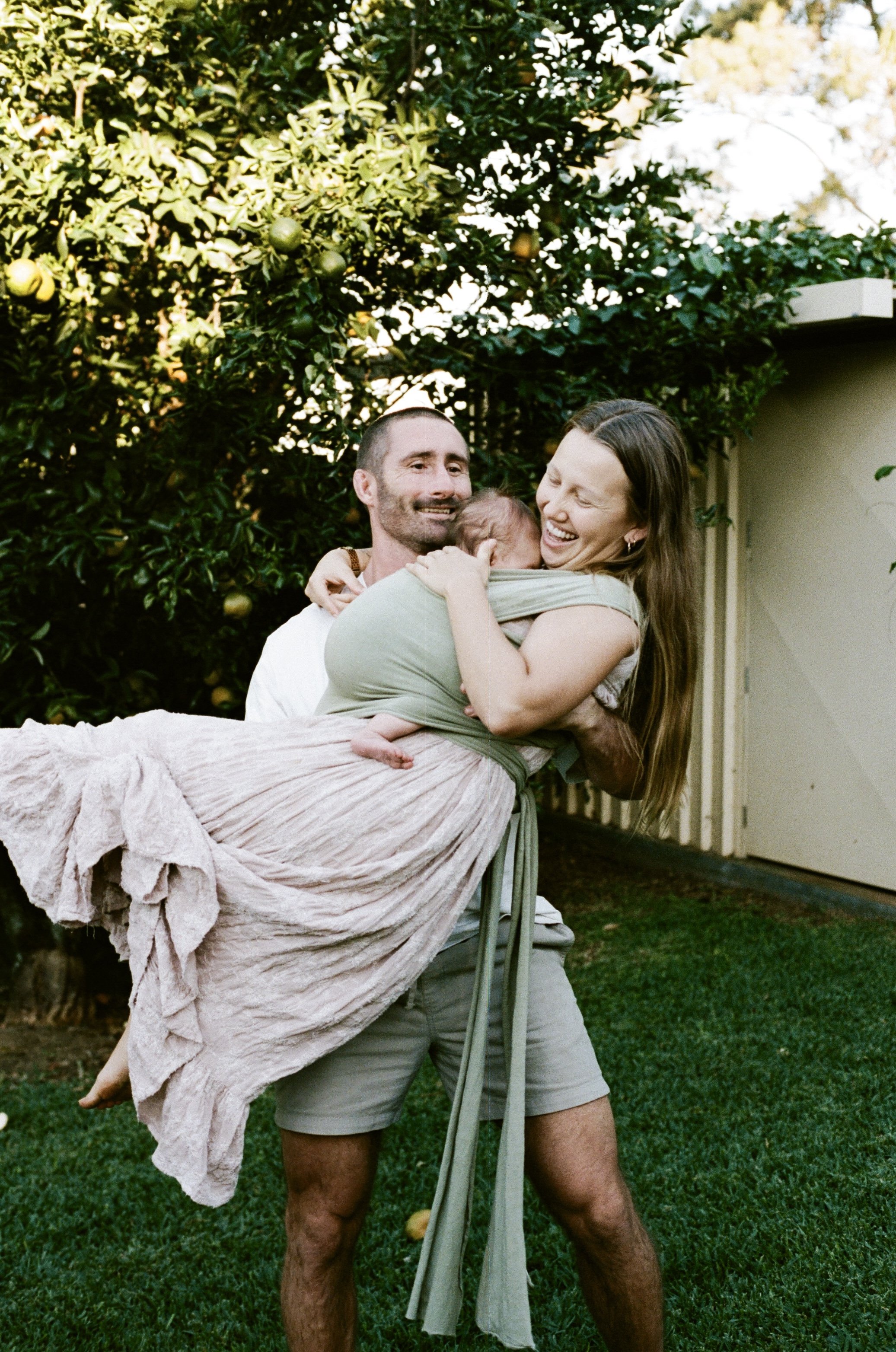 A man holding a woman and a baby outdoors near a green tree with fruit, during daytime.