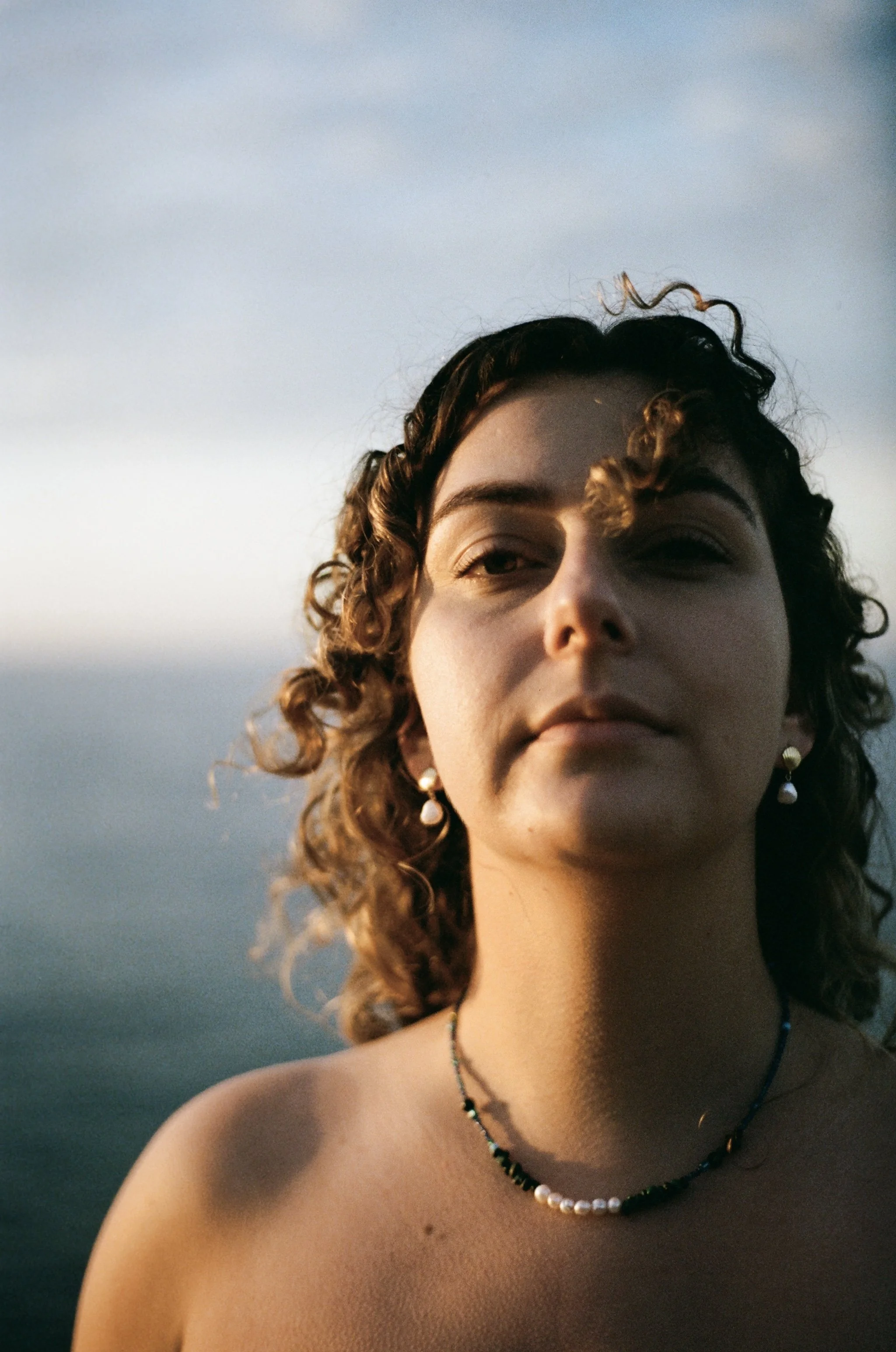A young woman with curly hair and earrings is taking a selfie outdoors during sunset, with a cloudy sky in the background.
