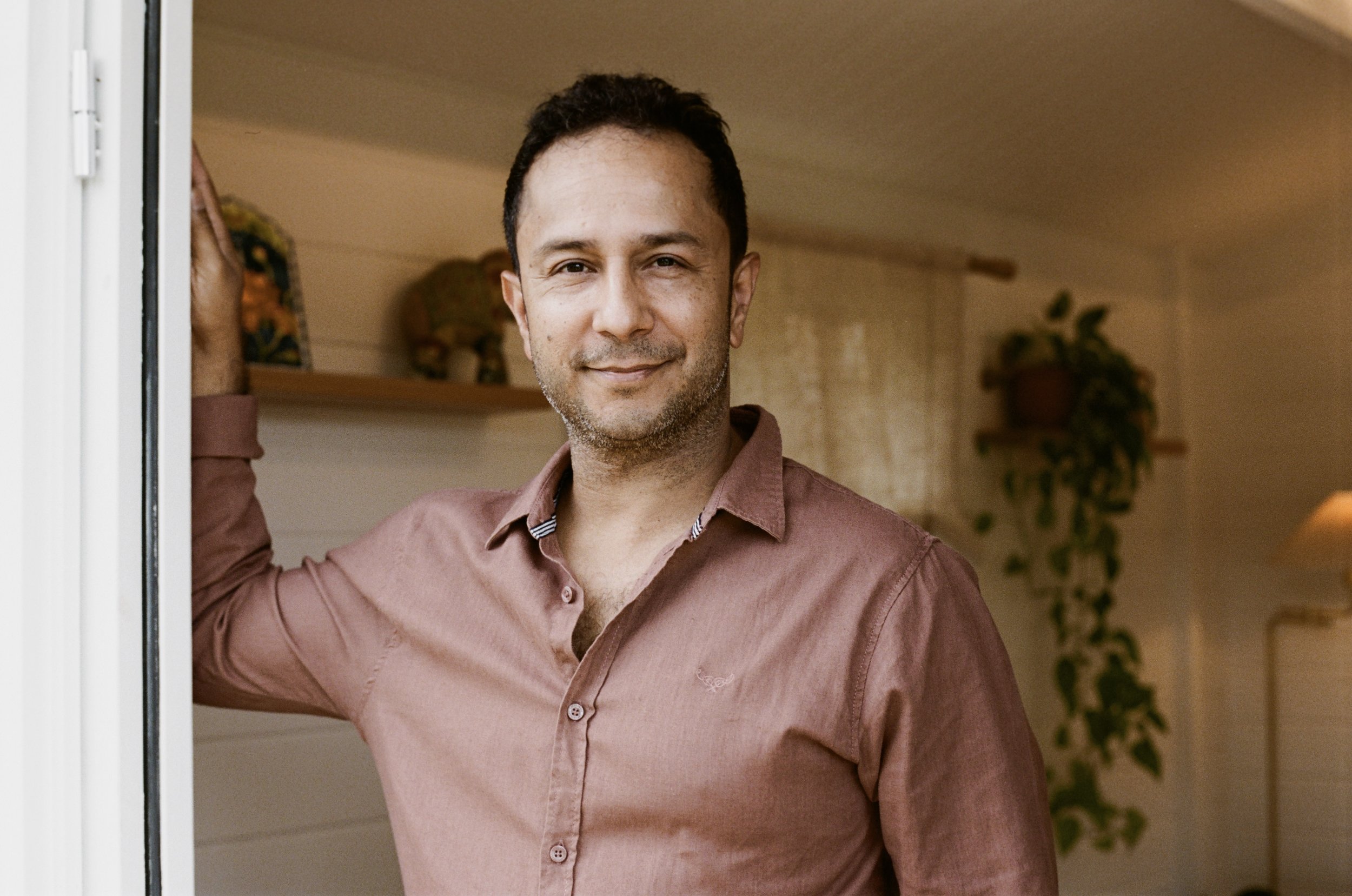 Man smiling and leaning on a doorframe in an indoor setting with plants in the background.