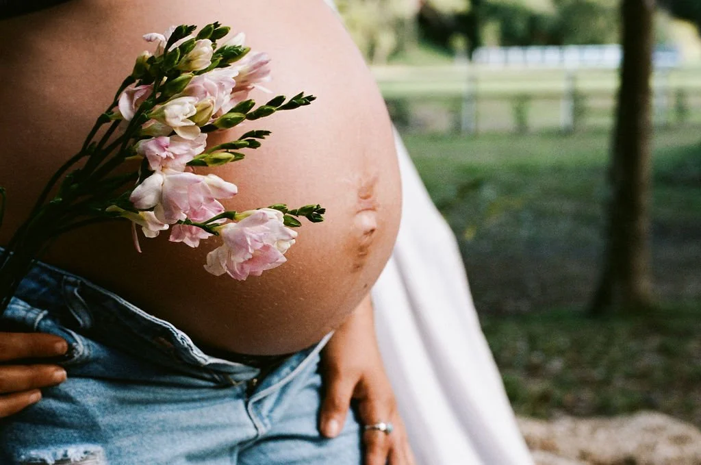 A pregnant woman holding a bouquet of pink flowers on her belly outdoors.