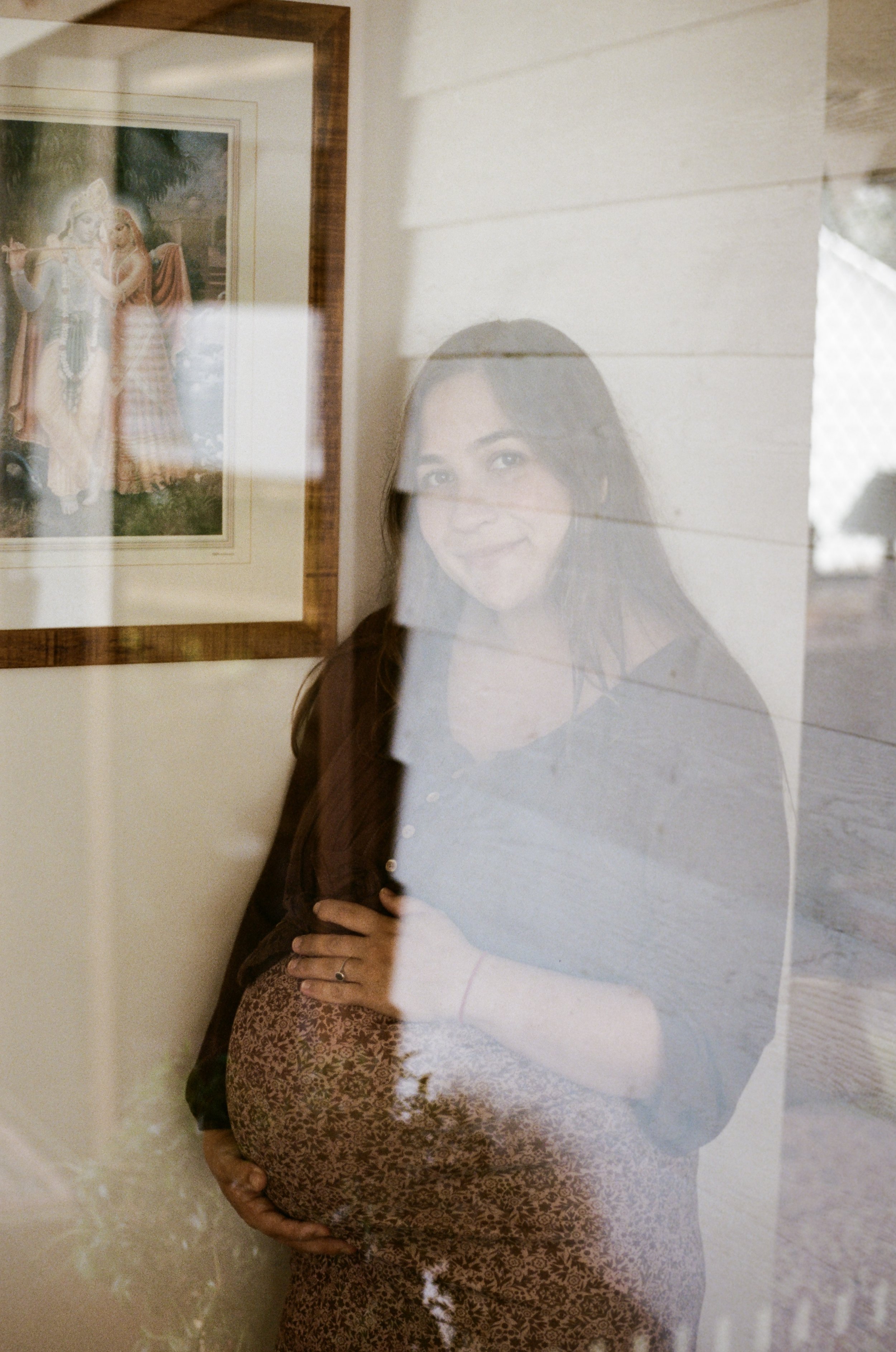 Reflection of a smiling woman with long hair, standing indoors against white wooden walls, with a framed painting of two figures playing flutes hanging on the wall behind her.