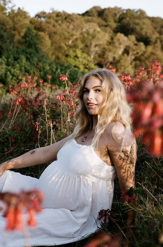 A pregnant woman with blonde hair wearing a white dress sitting among pink flowers in a lush outdoor setting with trees and hills in the background.