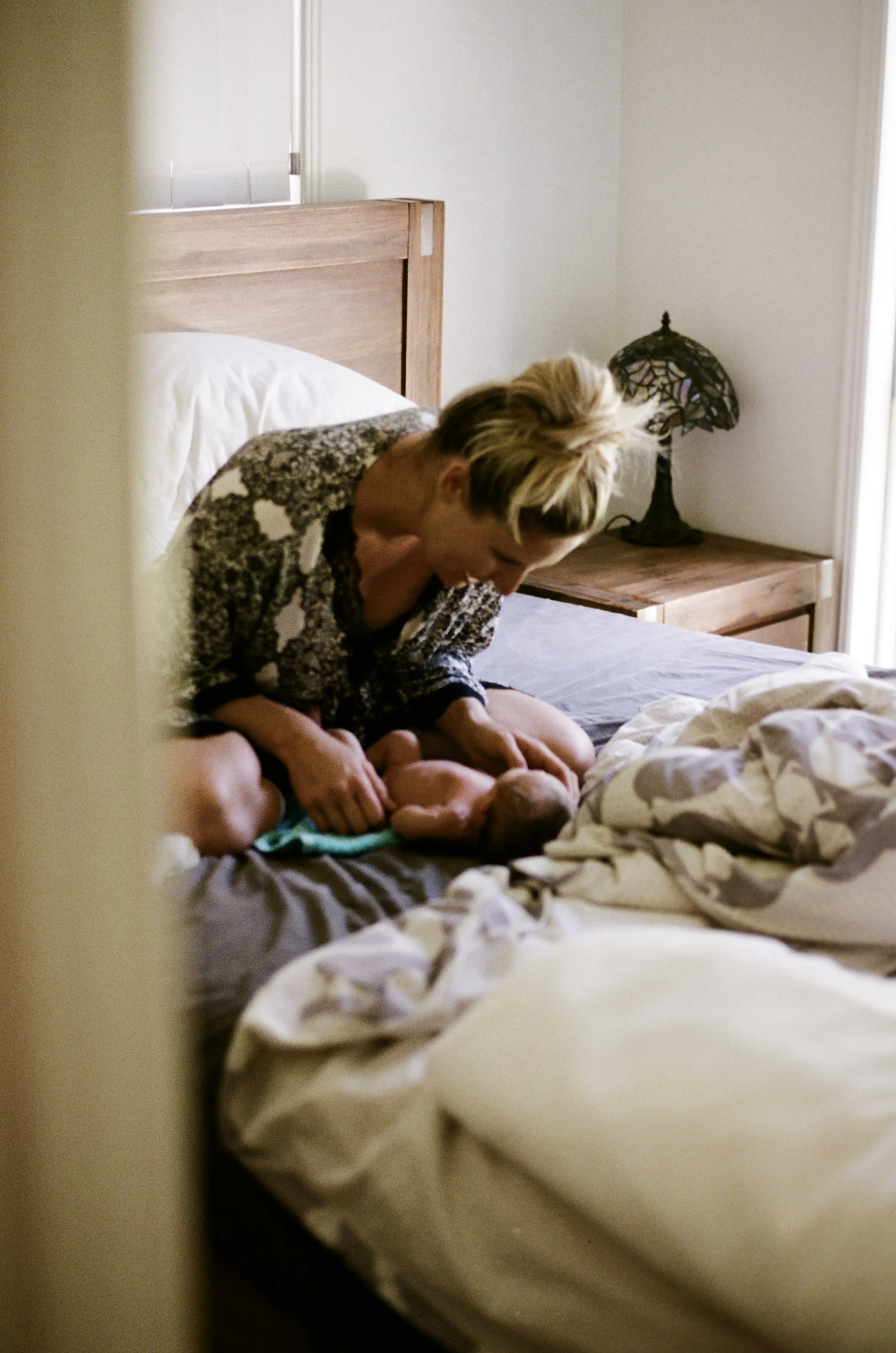 A woman leaning over a bed, breastfeeding a newborn baby. The bed has a wooden headboard, and there is a nightstand with a decorative lamp in the corner of the room. The photo is taken through a partially open door or a gap, giving a candid, intimate