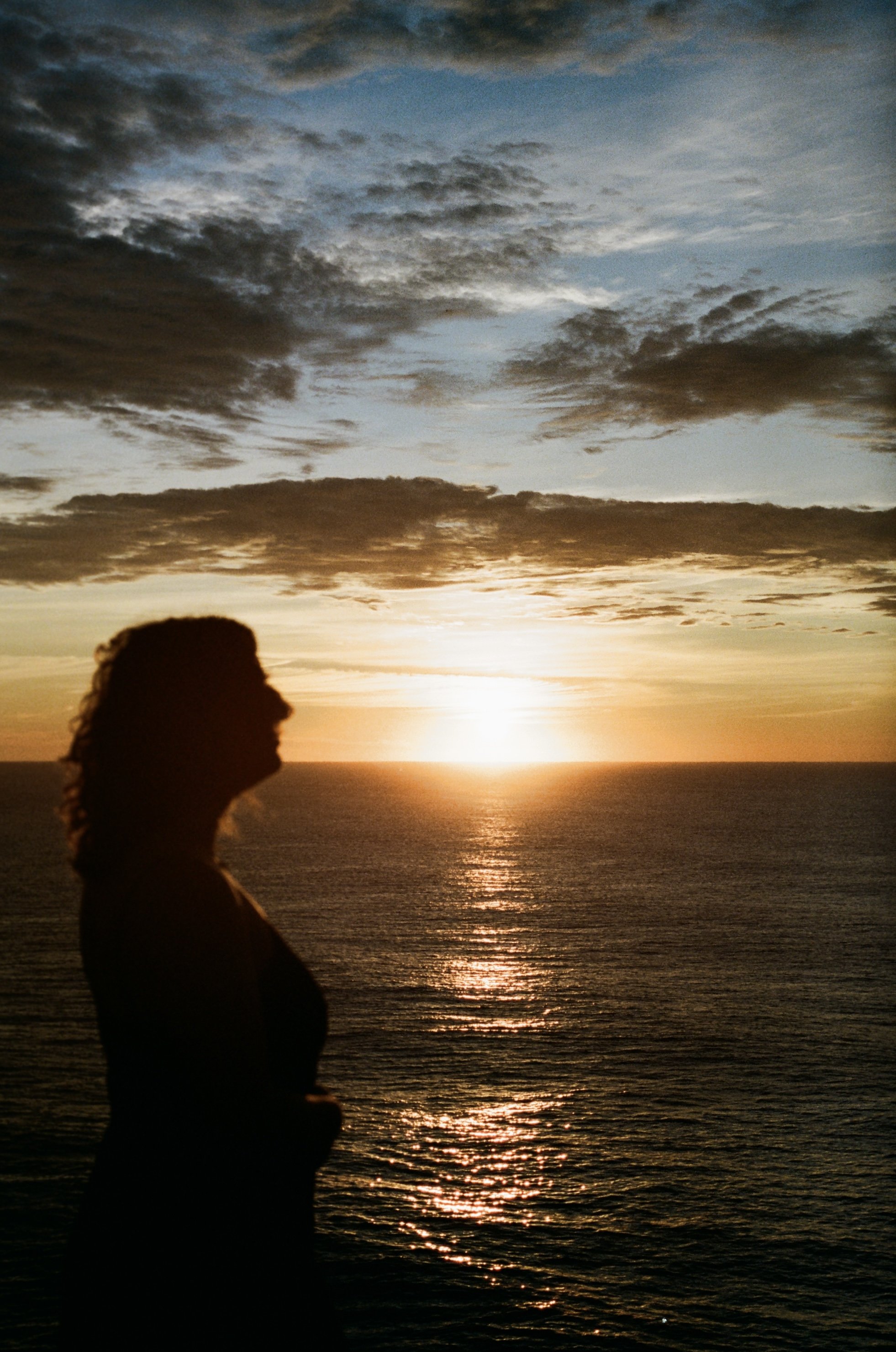 Silhouette of a person with curly hair standing by the water during sunset or sunrise with a cloudy sky.