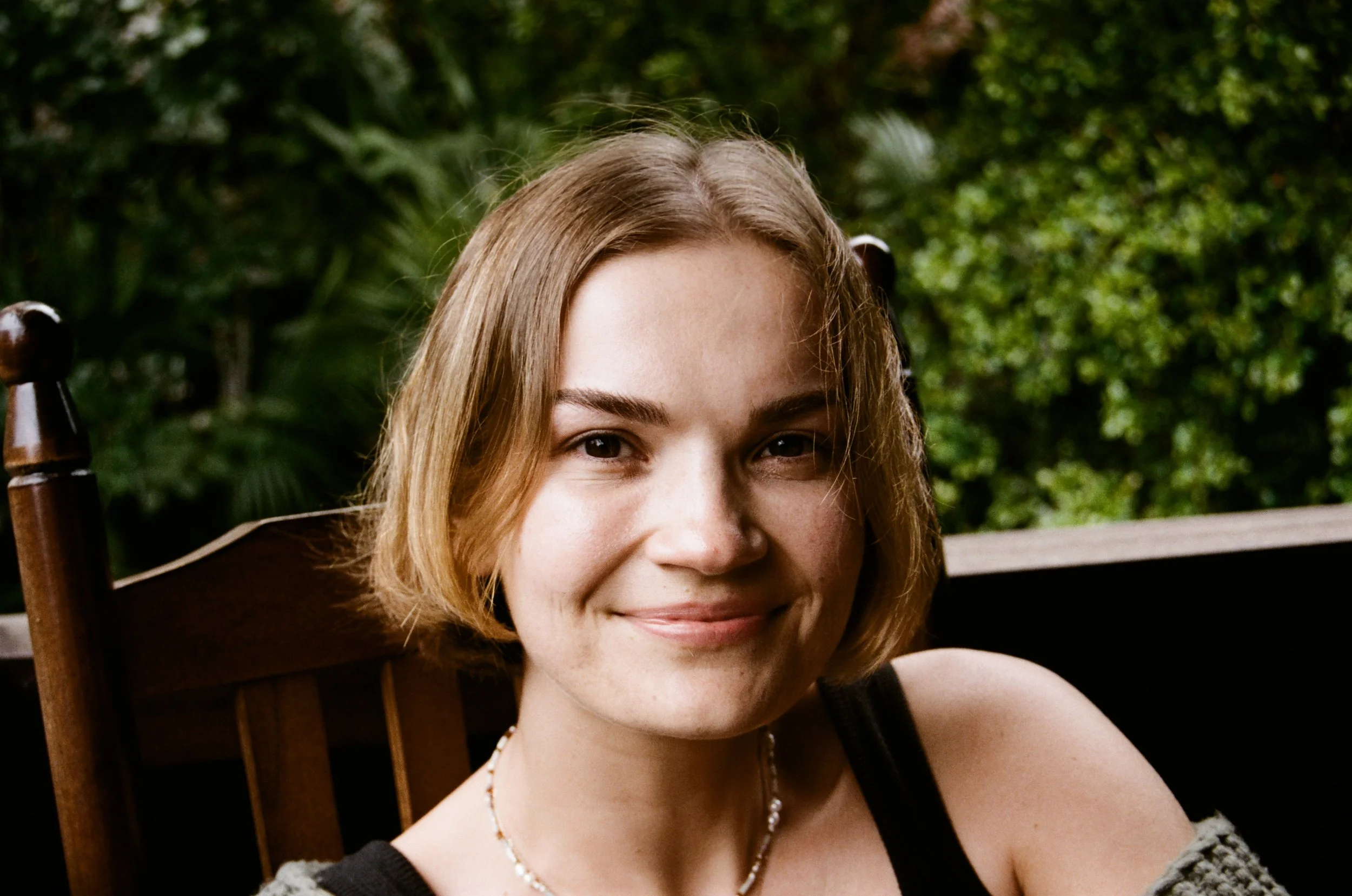 Smiling woman with blonde hair sitting outdoors with green foliage in the background.