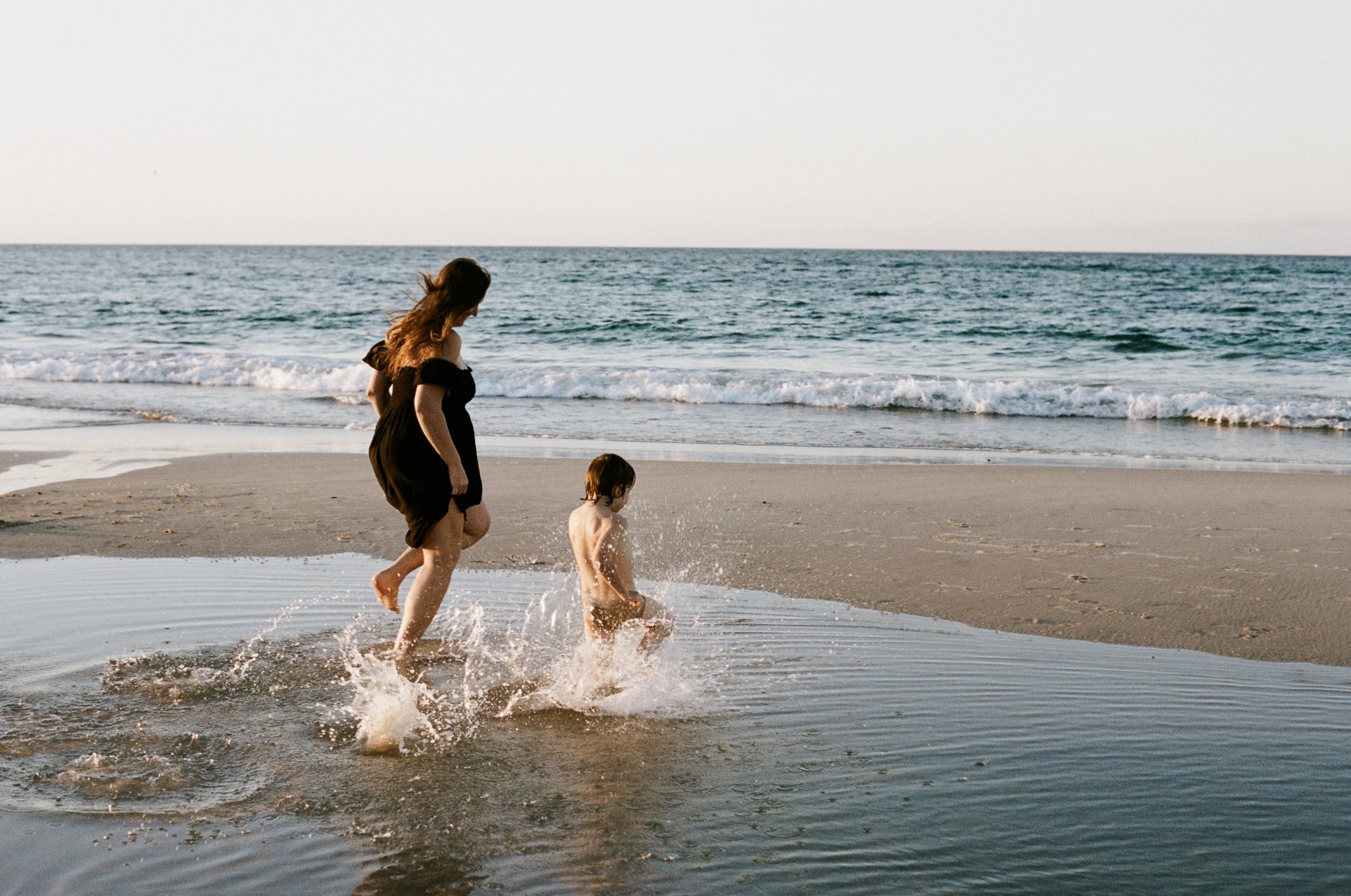 A woman and a young child playing and splashing in the shallow ocean water on a beach during daytime.