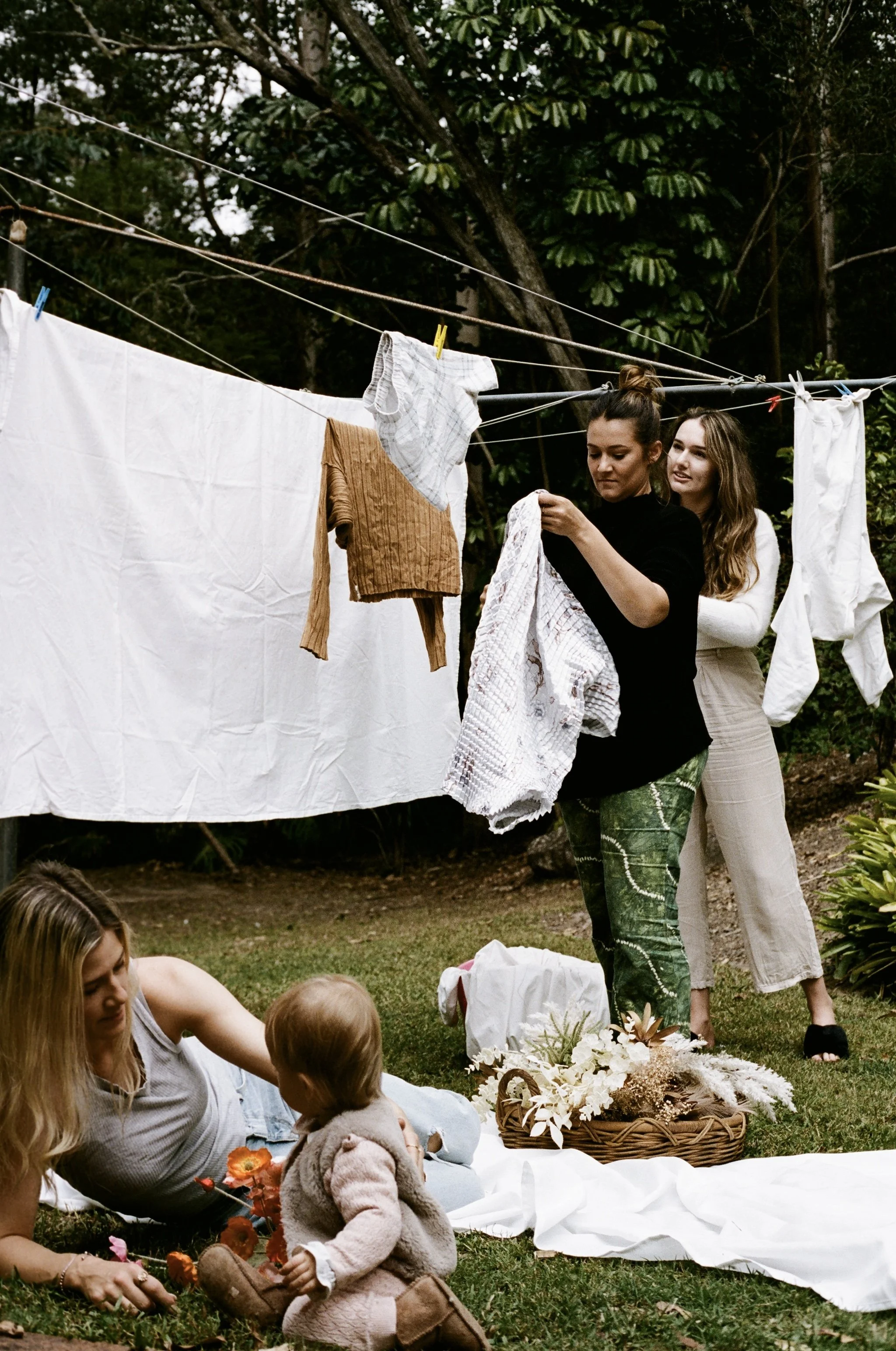 A woman and two children sitting on a white cloth on the grass, with a basket of flowers nearby, and another woman standing behind them hanging laundry on a clothesline outdoors surrounded by greenery.